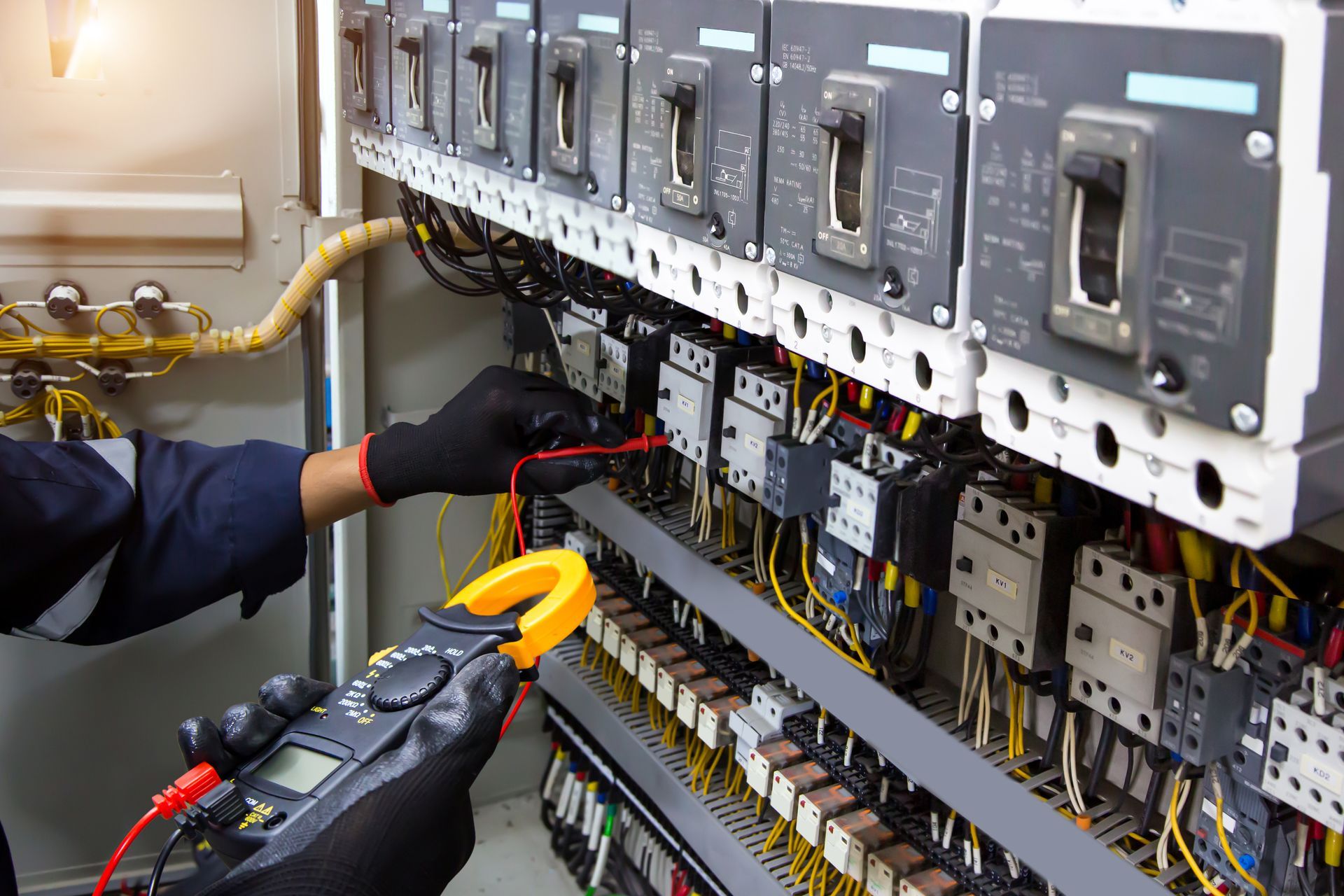 An electrician in black gloves uses a multimeter to test electrical components inside an industrial control panel.