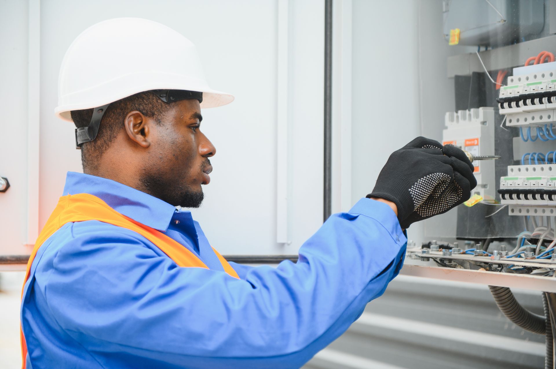 A technician in a hard hat and blue work shirt with an orange vest adjusts a switch inside an electrical circuit breaker.