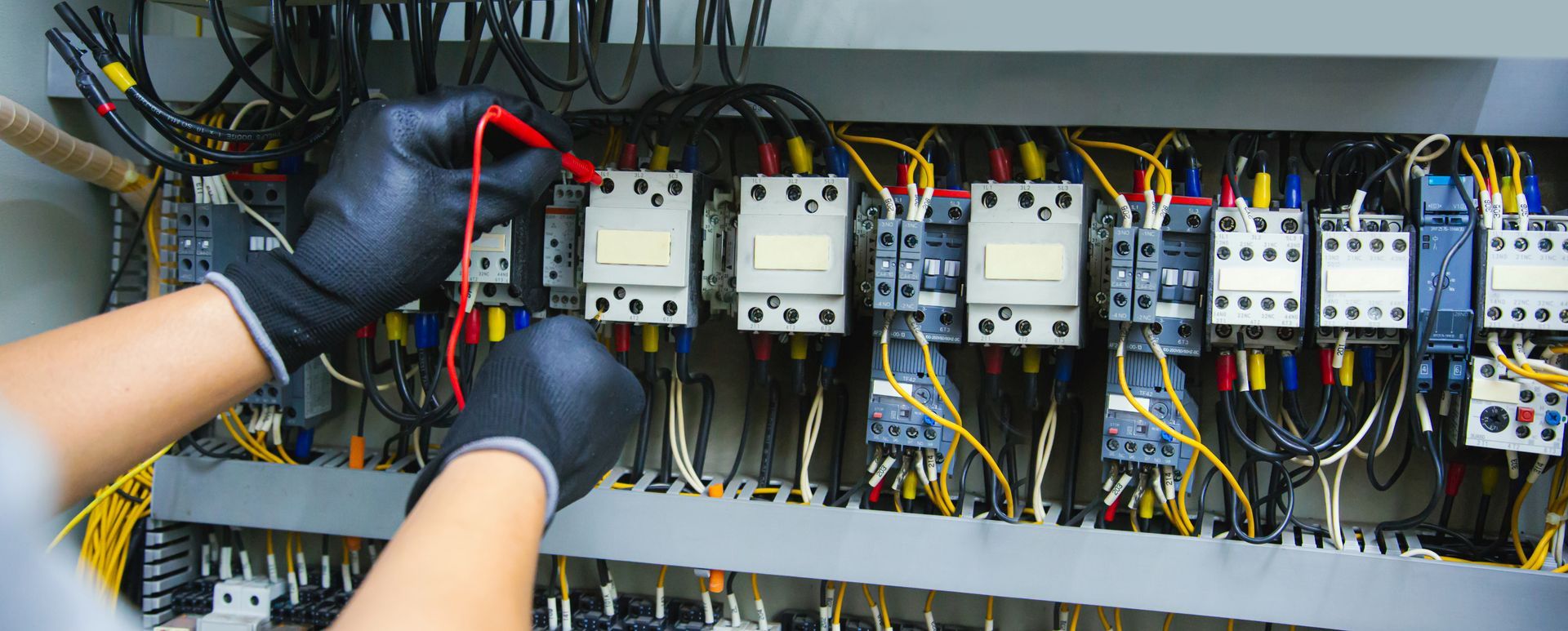 A technician wearing black gloves uses a multimeter probe to test electrical components in a control panel.