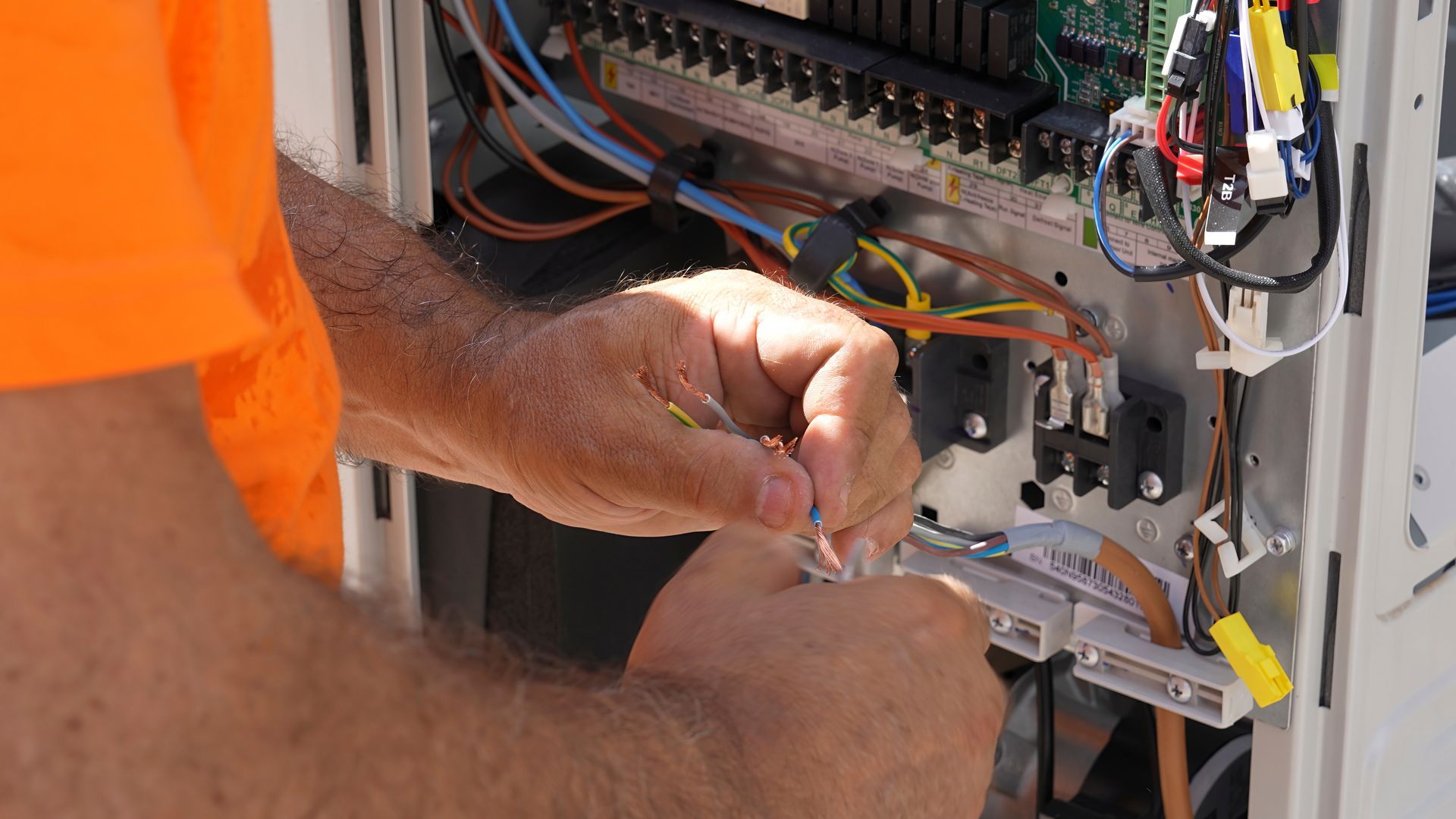 Technician in an orange shirt wires electrical components inside an equipment cabinet.