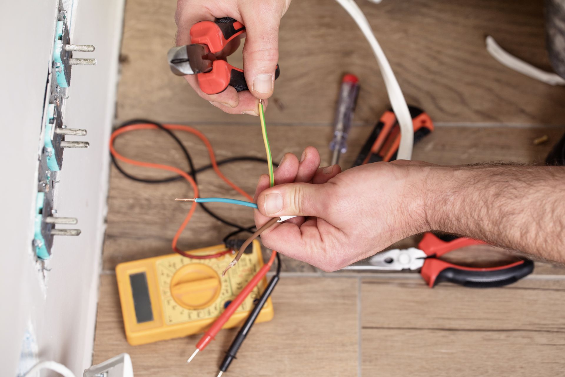 A person uses wire cutters on a green electrical wire, with other tools and a multimeter nearby on a wooden surface.