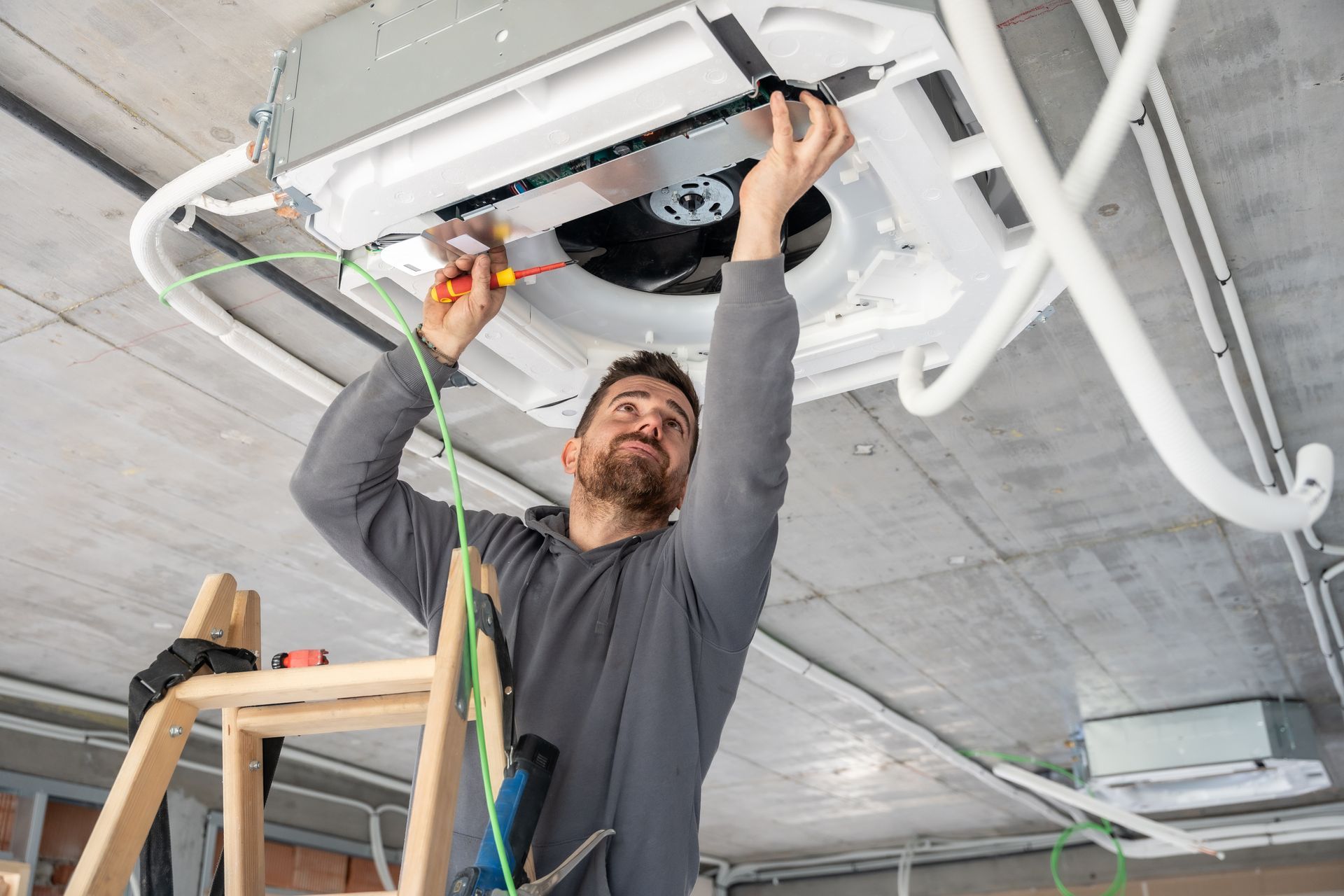 A person standing on a ladder, installing or repairing a ceiling-mounted HVAC unit in an unfinished room.