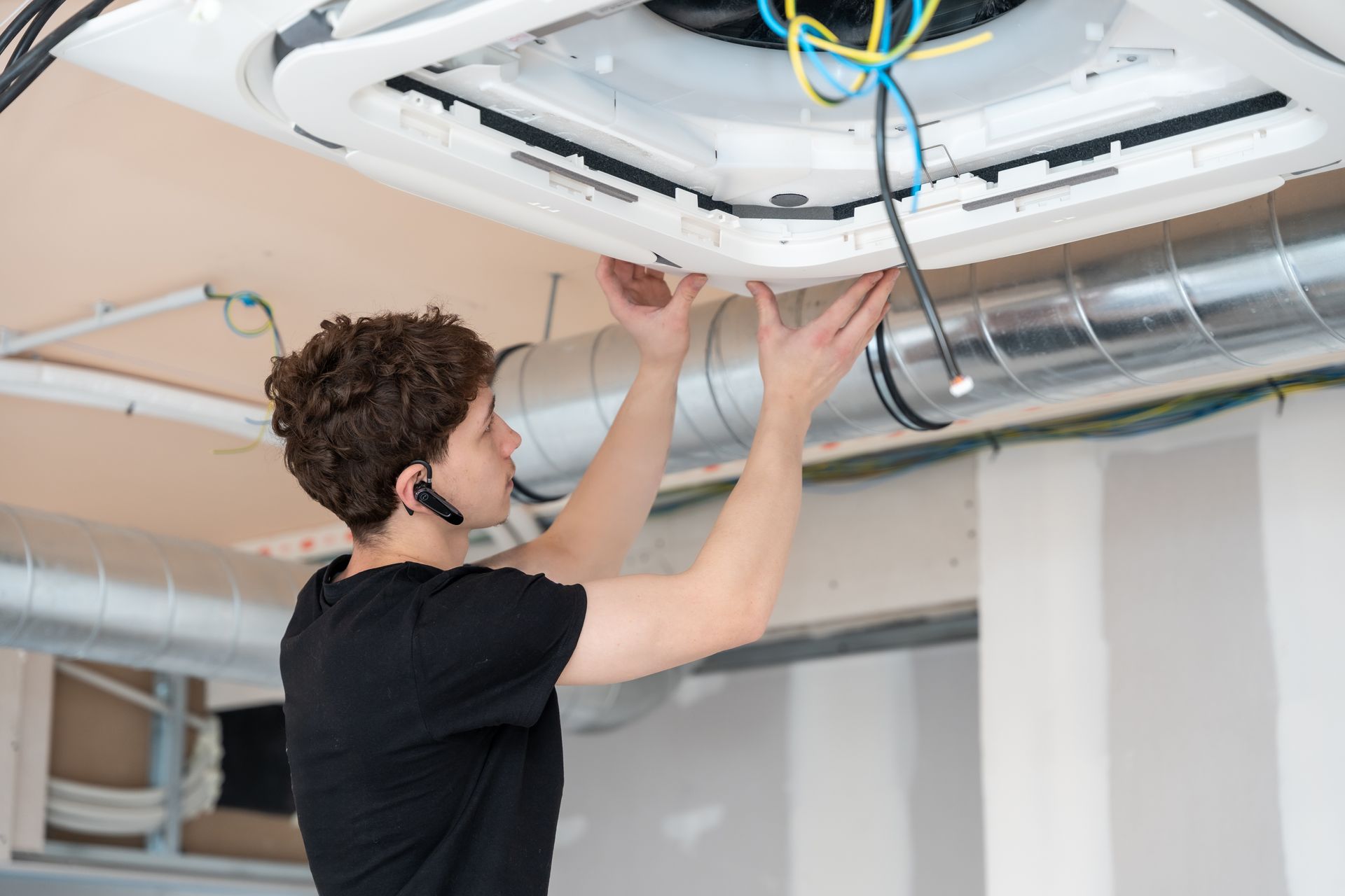 A person in a black shirt installs a white ceiling-mounted HVAC unit in an unfinished room with metal ductwork.