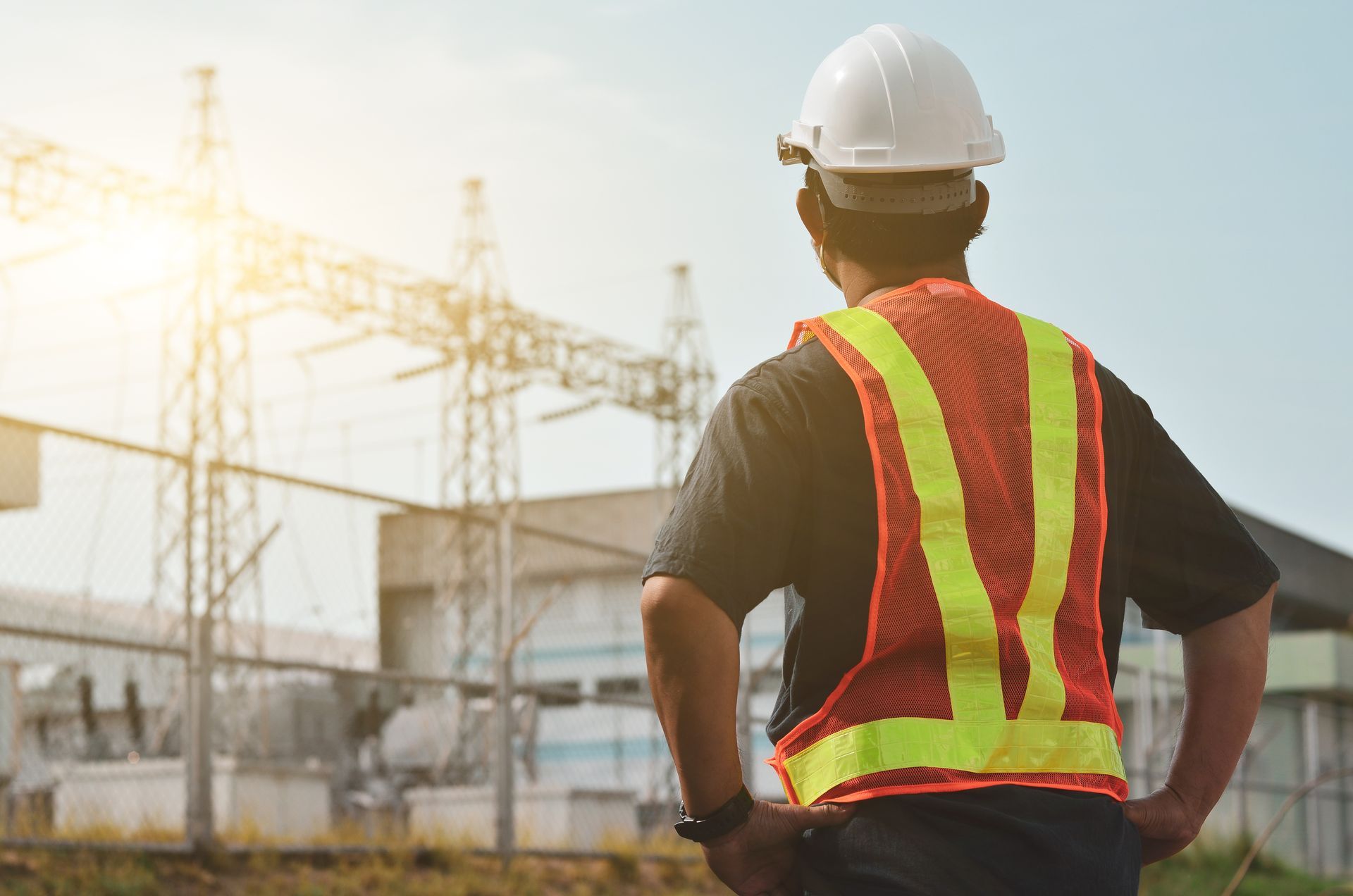 A person in a hard hat and high-visibility vest stands with their back to the camera, looking at an electrical substation.