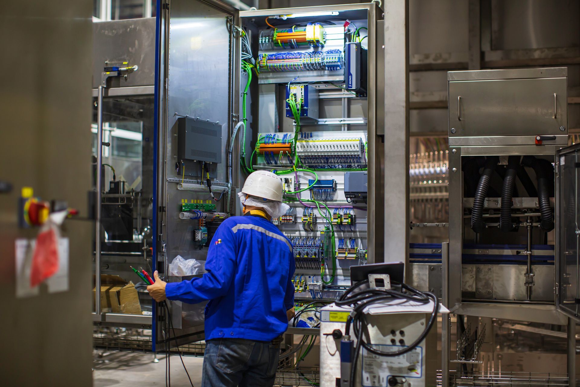 A person in a blue uniform and hard hat working on the open wiring and electrical components of an industrial machine.