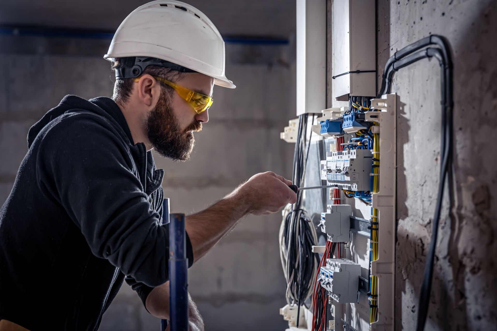 A person wearing a white hard hat and safety glasses uses a screwdriver on an open electrical panel on a wall.