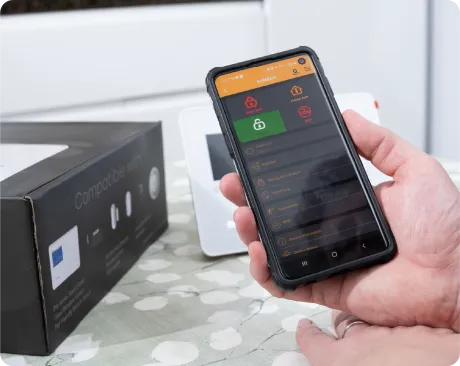 Person holding phone showing home security app, black box, and white security panel in background.