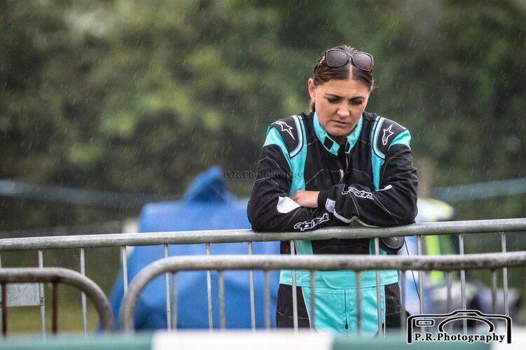 A woman in a race suit is leaning on a fence.