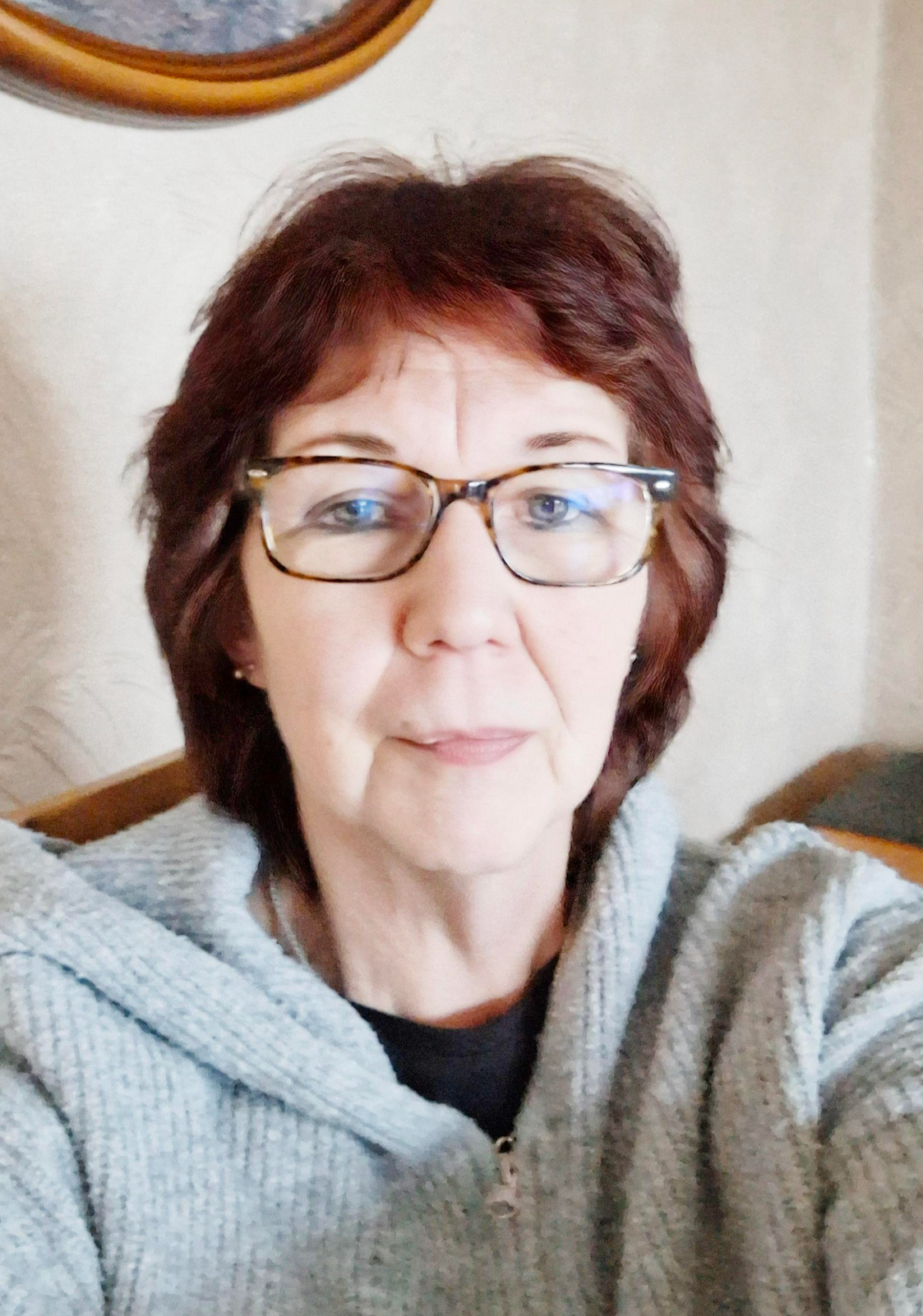 Woman with auburn hair and glasses smiles. Wearing a gray zip-up sweater.