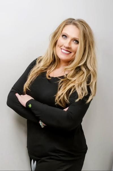 Smiling blonde woman in a black outfit standing with arms crossed against a light gray wall
