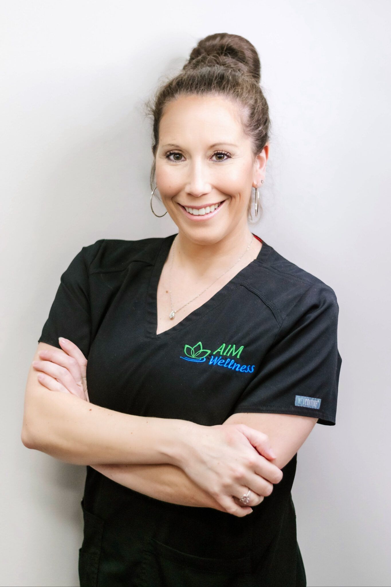 Smiling healthcare worker in black scrubs with arms crossed against a light gray background