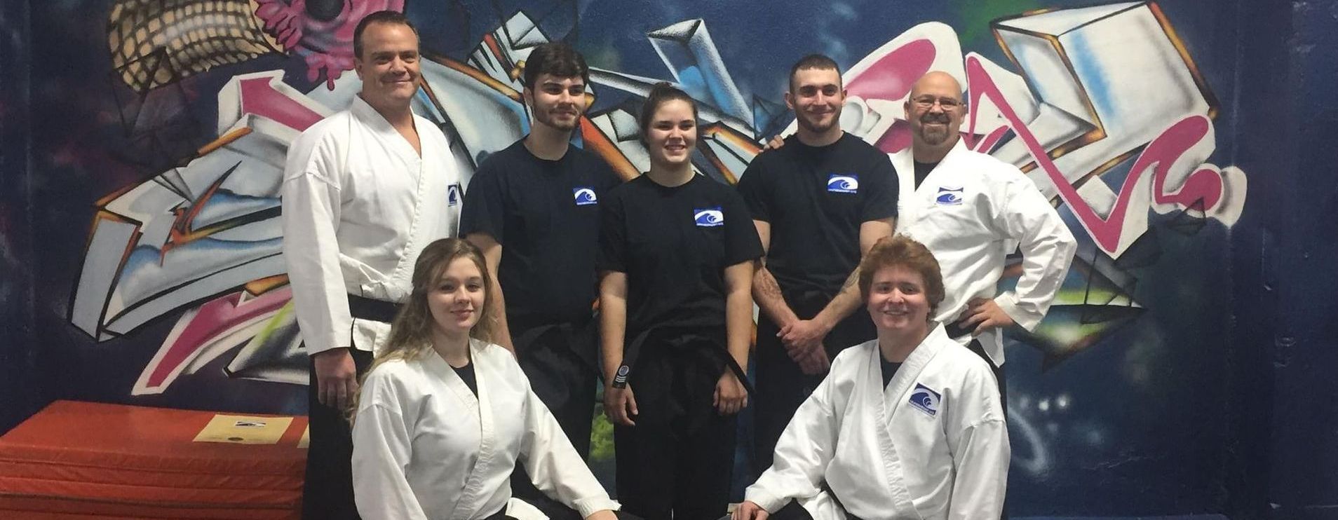 Group of people, some in martial arts uniforms, pose in front of a graffiti mural.