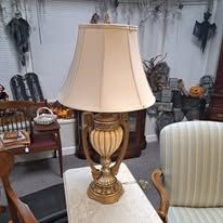 A vintage gold table lamp with a white shade on a table. Dark background with spooky decorations.