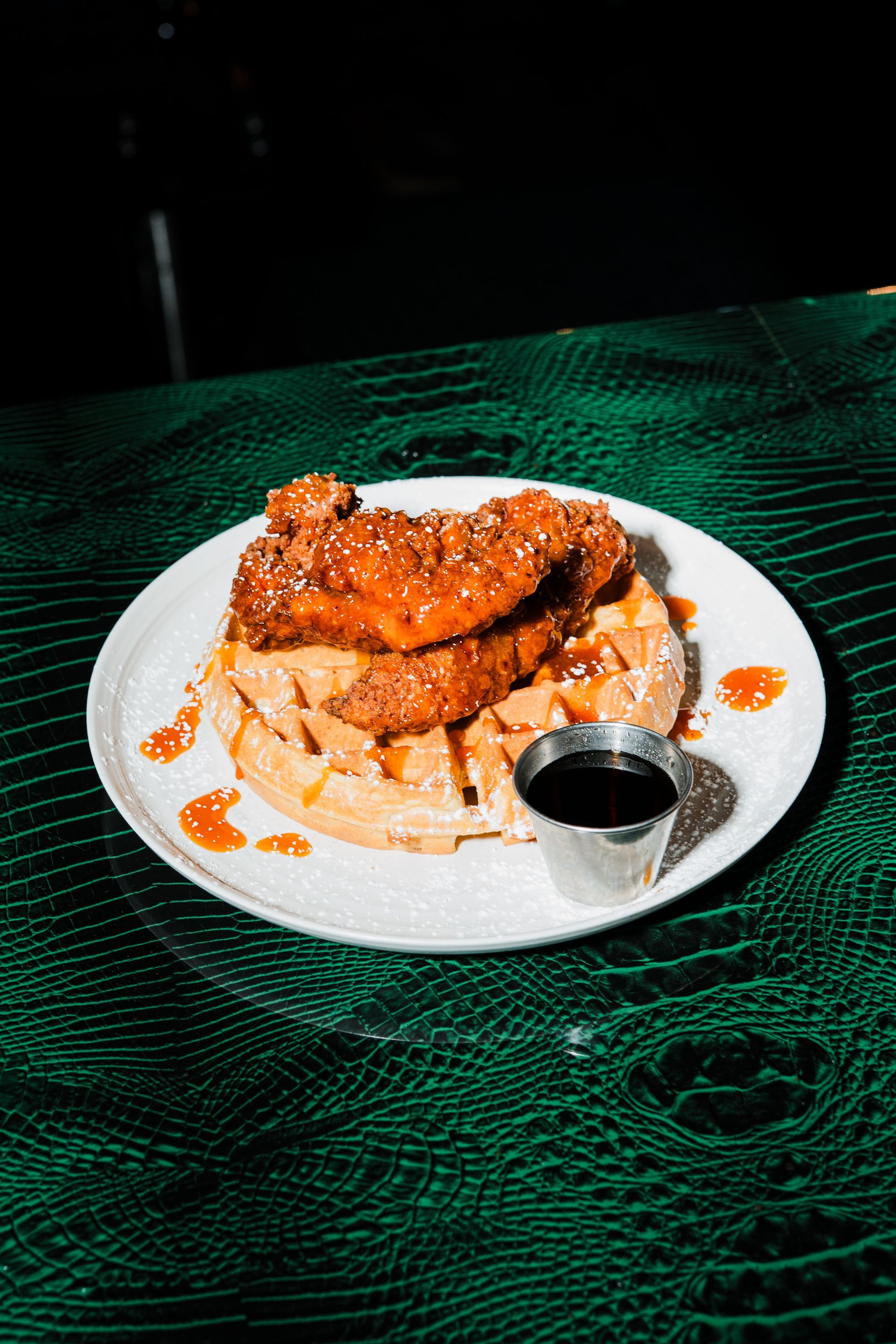 Chicken and waffles with syrup on a white plate, served on a textured green surface.