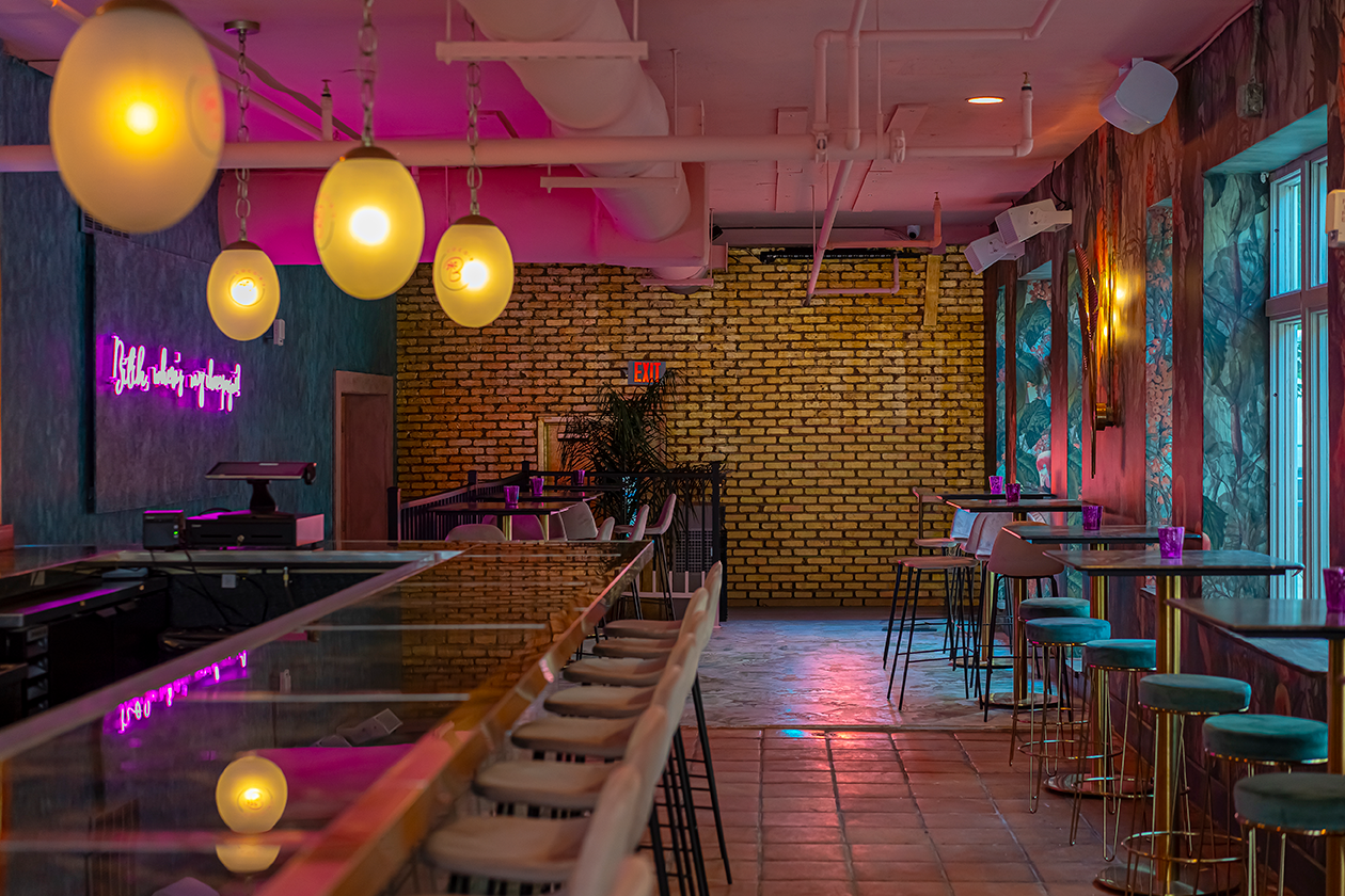 Interior view of a bar with a long counter, stools, gold wall, and hanging lights. Pink and yellow lighting.