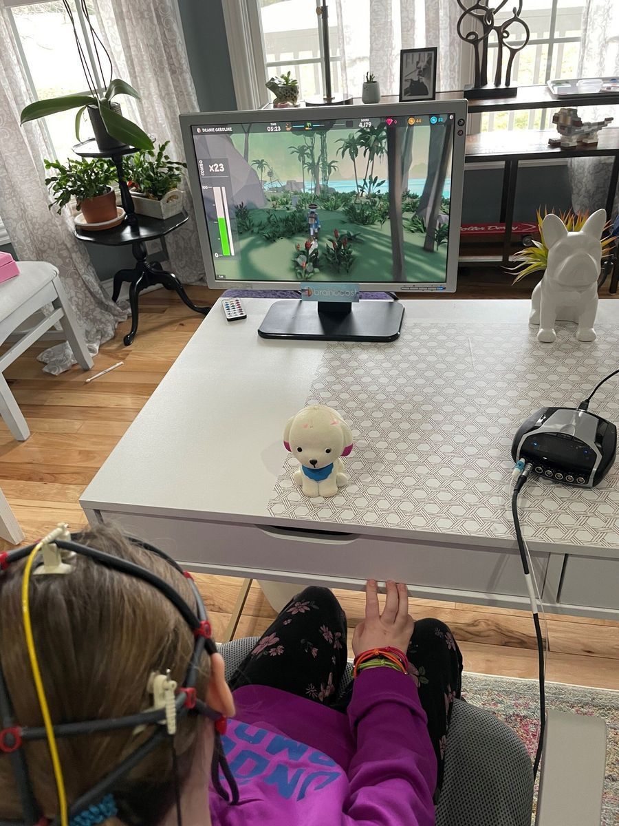 Child wearing a head sensor plays a video game on a desktop monitor at a white table. A dog figurine sits on the table.