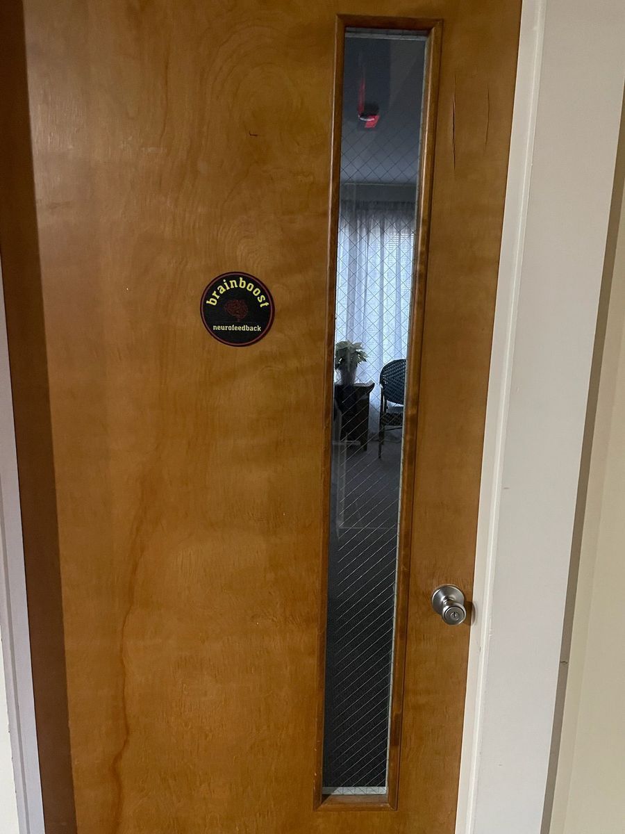 A wooden door with a long vertical window and a round name plaque. The window reveals a room with chairs and curtains.