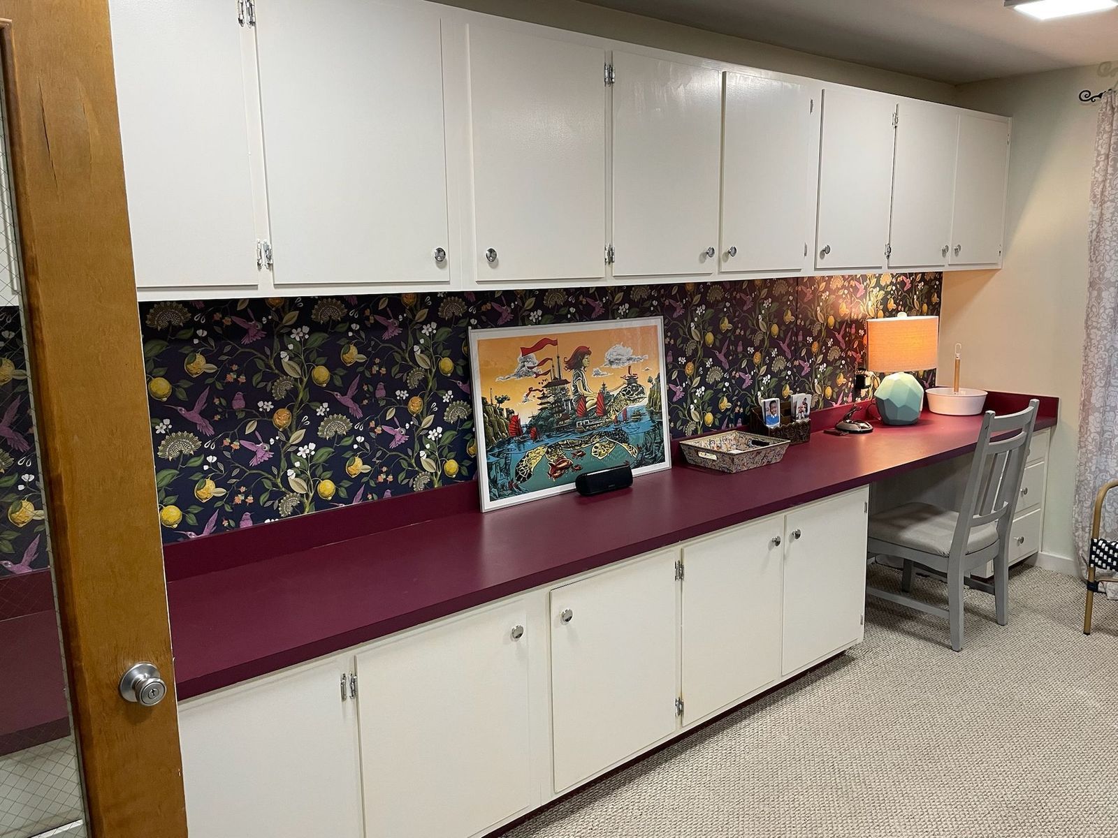 White cabinets above and below a desk with a magenta surface.  Floral wallpaper fills the space between. A framed picture sits on the desk.