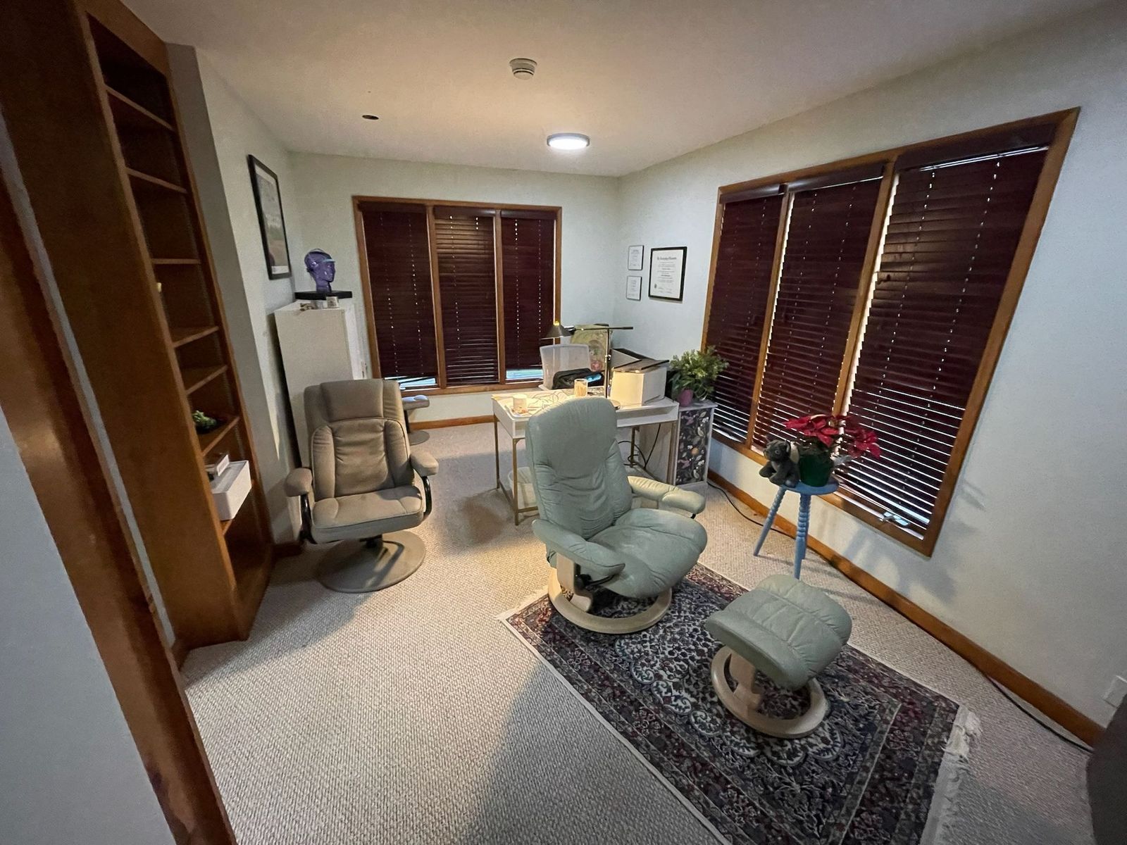 A light-filled office with a patterned rug, recliner chairs, and a desk by shuttered windows. A bookcase lines one wall.