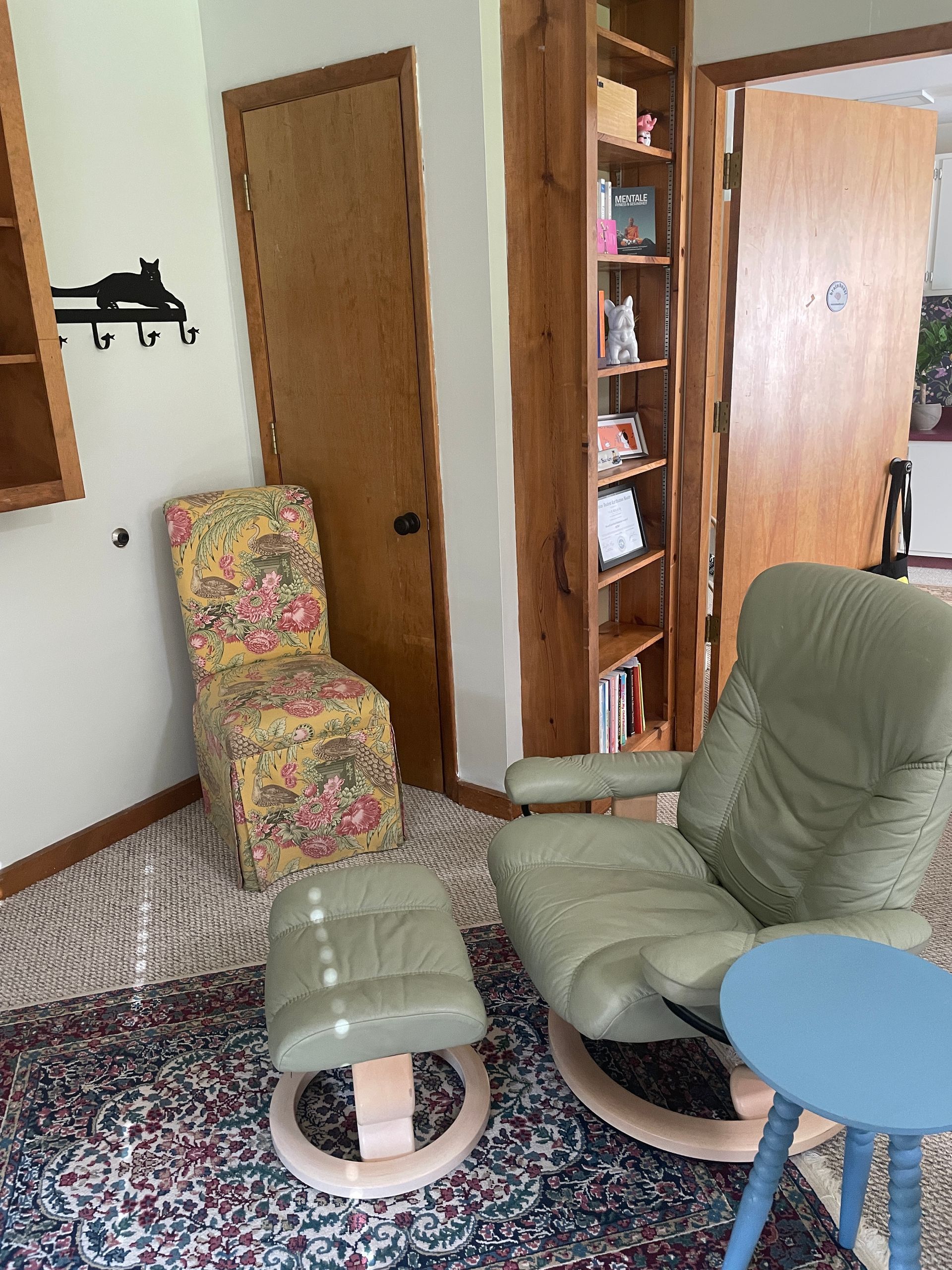 A room with a floral chair, recliner with ottoman, and a bookshelf by a doorway. A blue side table is nearby.