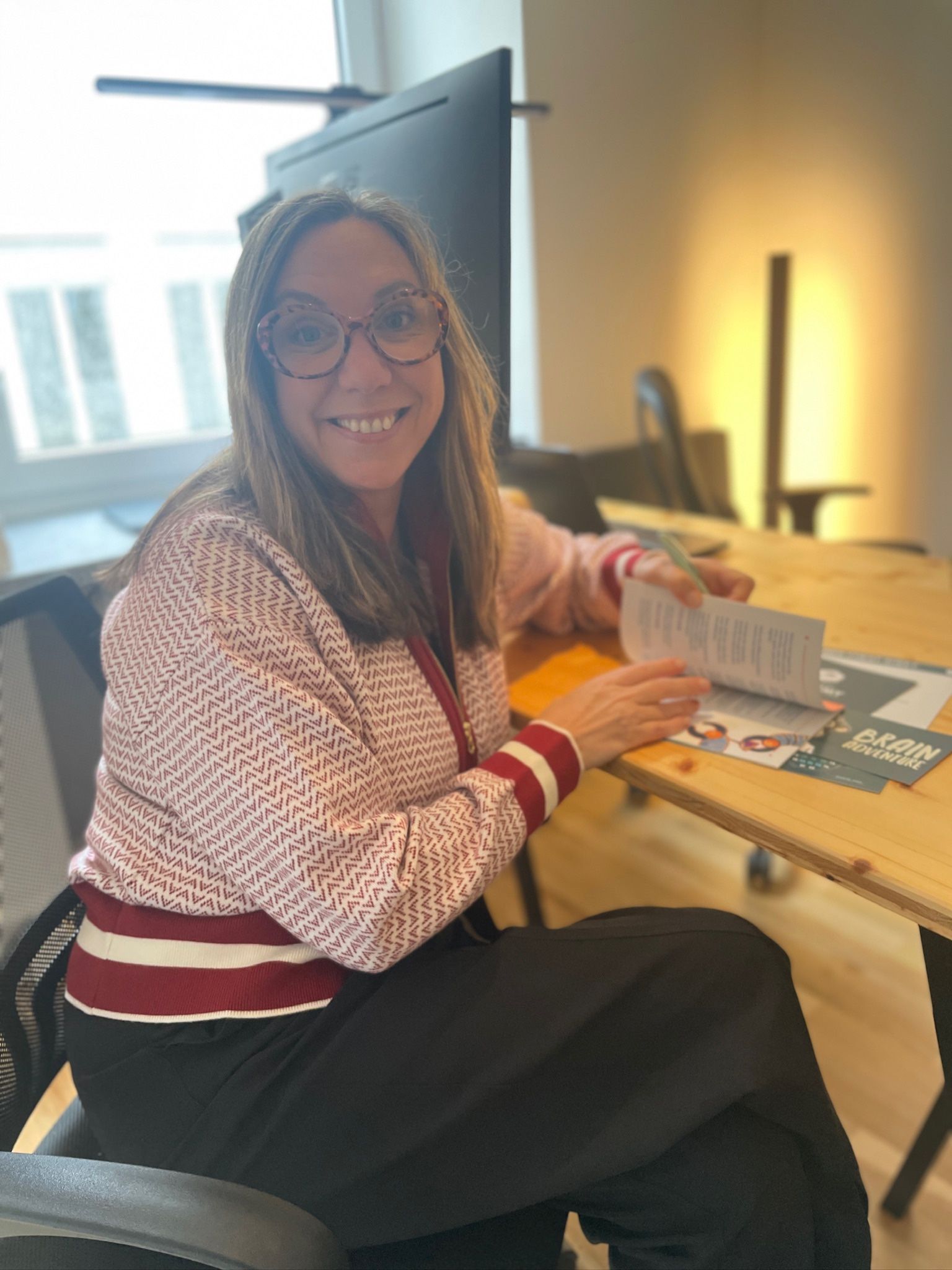 Woman wearing glasses and a pink jacket sits at a desk, smiling and looking at a pamphlet. She is in an office setting.