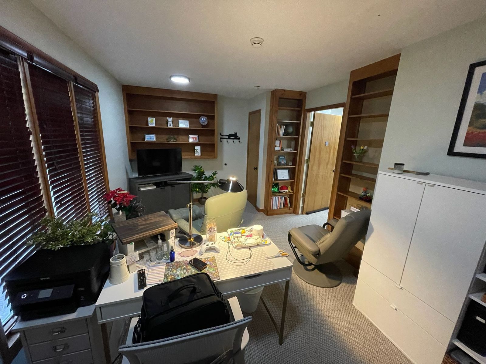 A cluttered home office with desk, shelves, and window with blinds. Features a printer, computer screen, and a swivel chair.