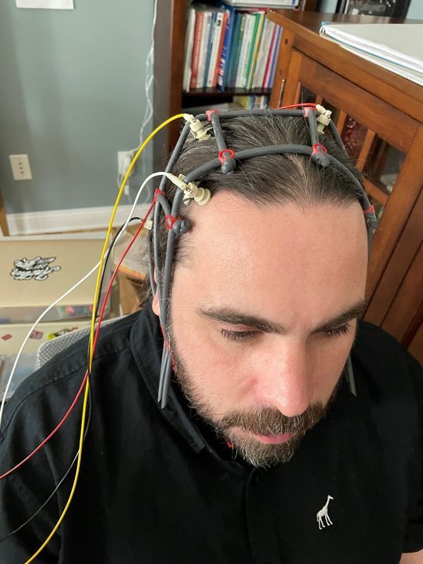 Man with dark hair and beard wearing a head-mounted device with wires. The device is likely for brain imaging.