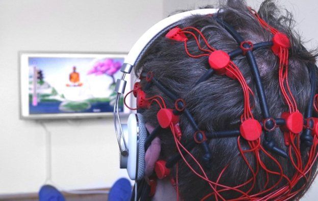 Person wearing a headset with red sensors connected to a device, viewing a screen with a brain image; indoors.