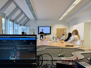 A presentation in a conference room: a man standing near a screen, people seated at a table, and a laptop with data visible.