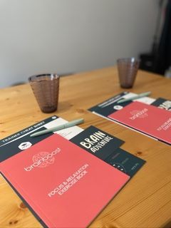 Two Brainboost workbooks and glasses sit on a wooden table, with a pen.