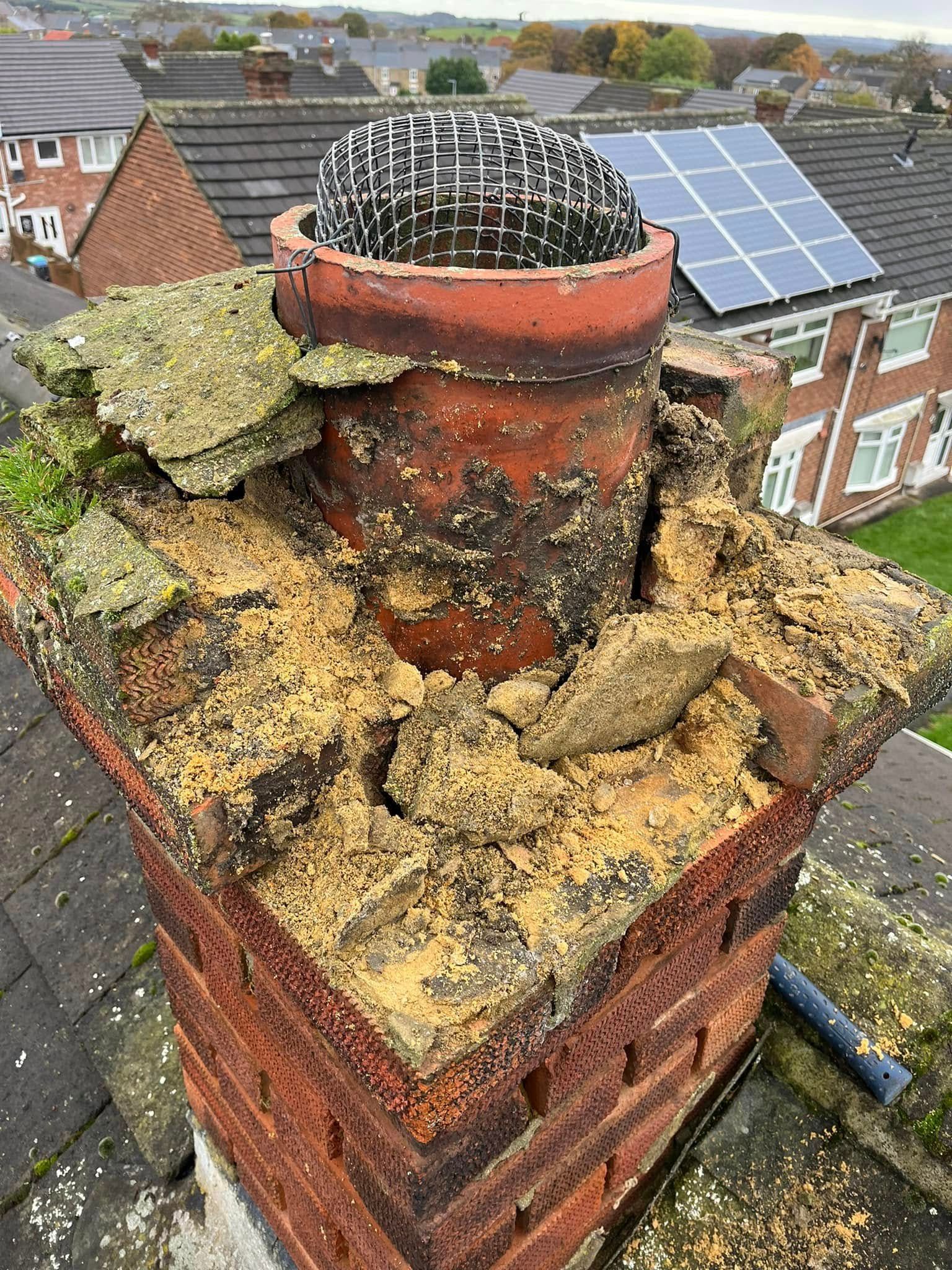 A red brick chimney rises from the weathered, damaged roof of a house against a clear blue sky.
