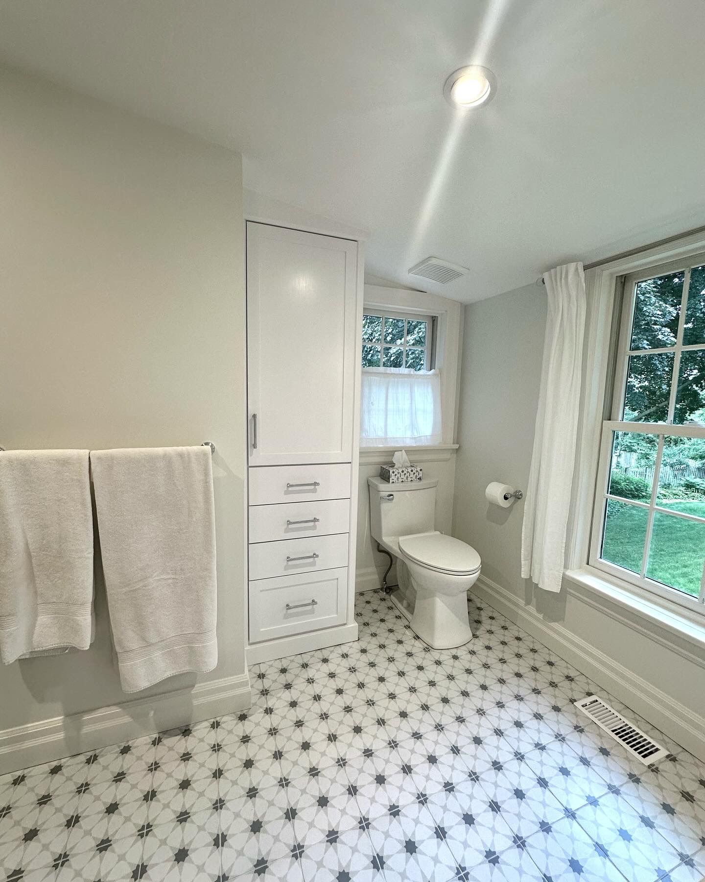 Bathroom with white cabinetry, toilet, and patterned tile floor. Towel rack on the left, window with view on right.