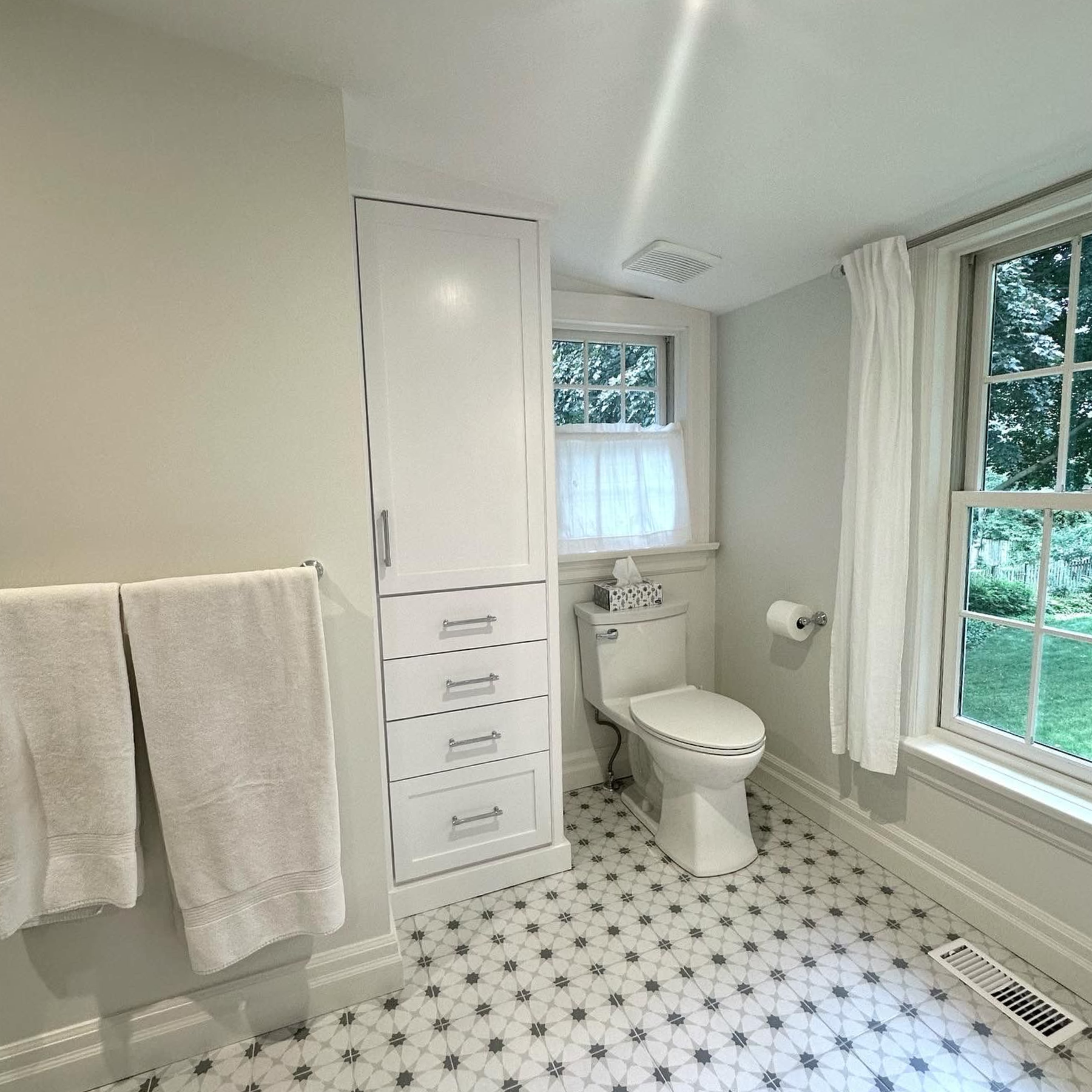 Bathroom with white cabinetry, toilet, patterned floor, and two windows with sheer curtains.