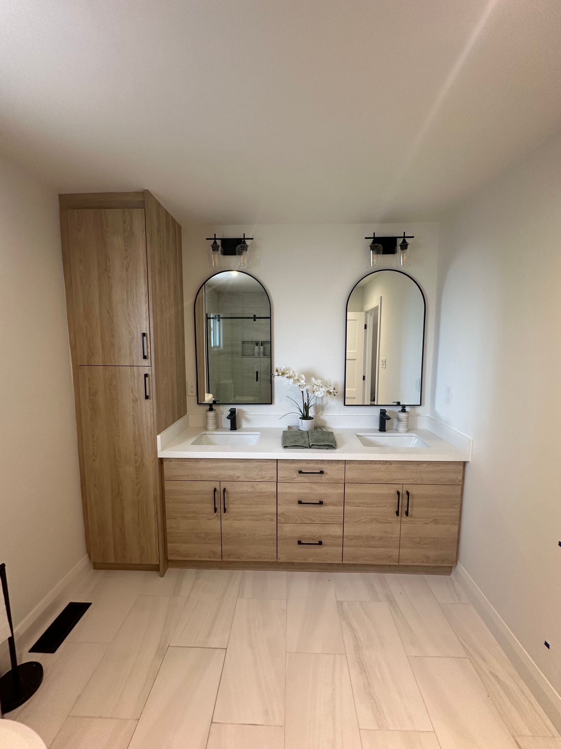 Bathroom with double vanity, arched mirrors, light wood cabinets, and marble-like countertops.