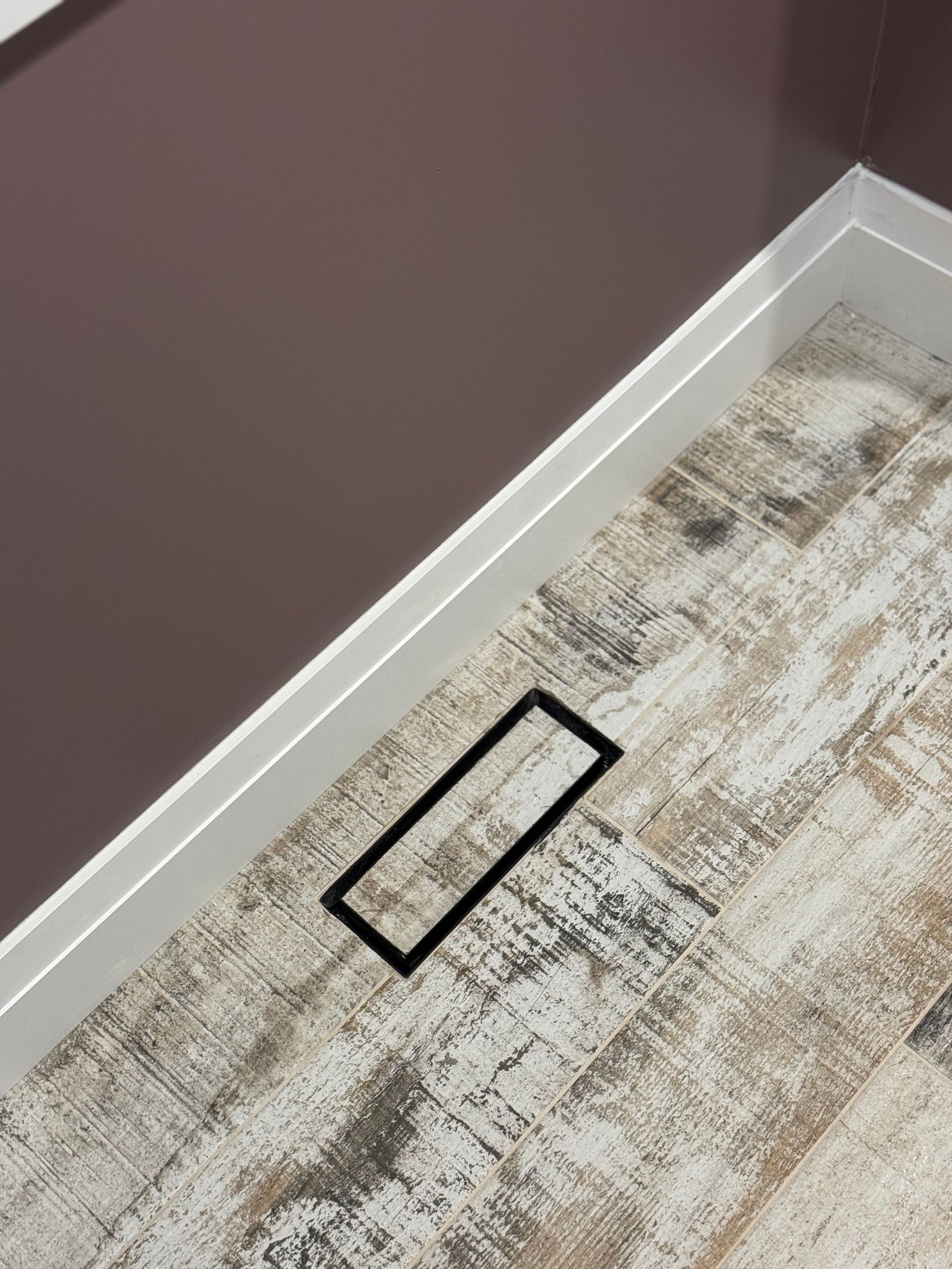 Rectangular black drain in a shower with wood-look flooring and dark mauve wall.