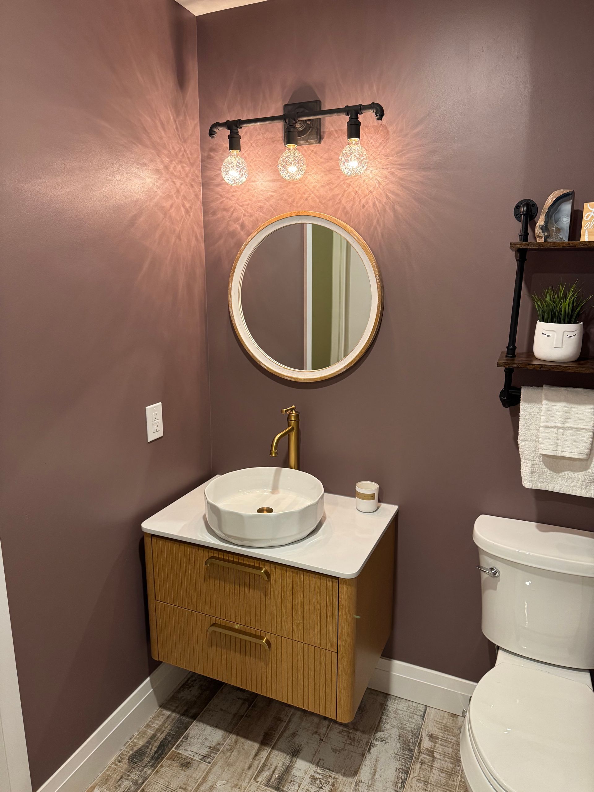 Small bathroom with floating wooden vanity, gold faucet, round mirror, and purple-mauve walls.