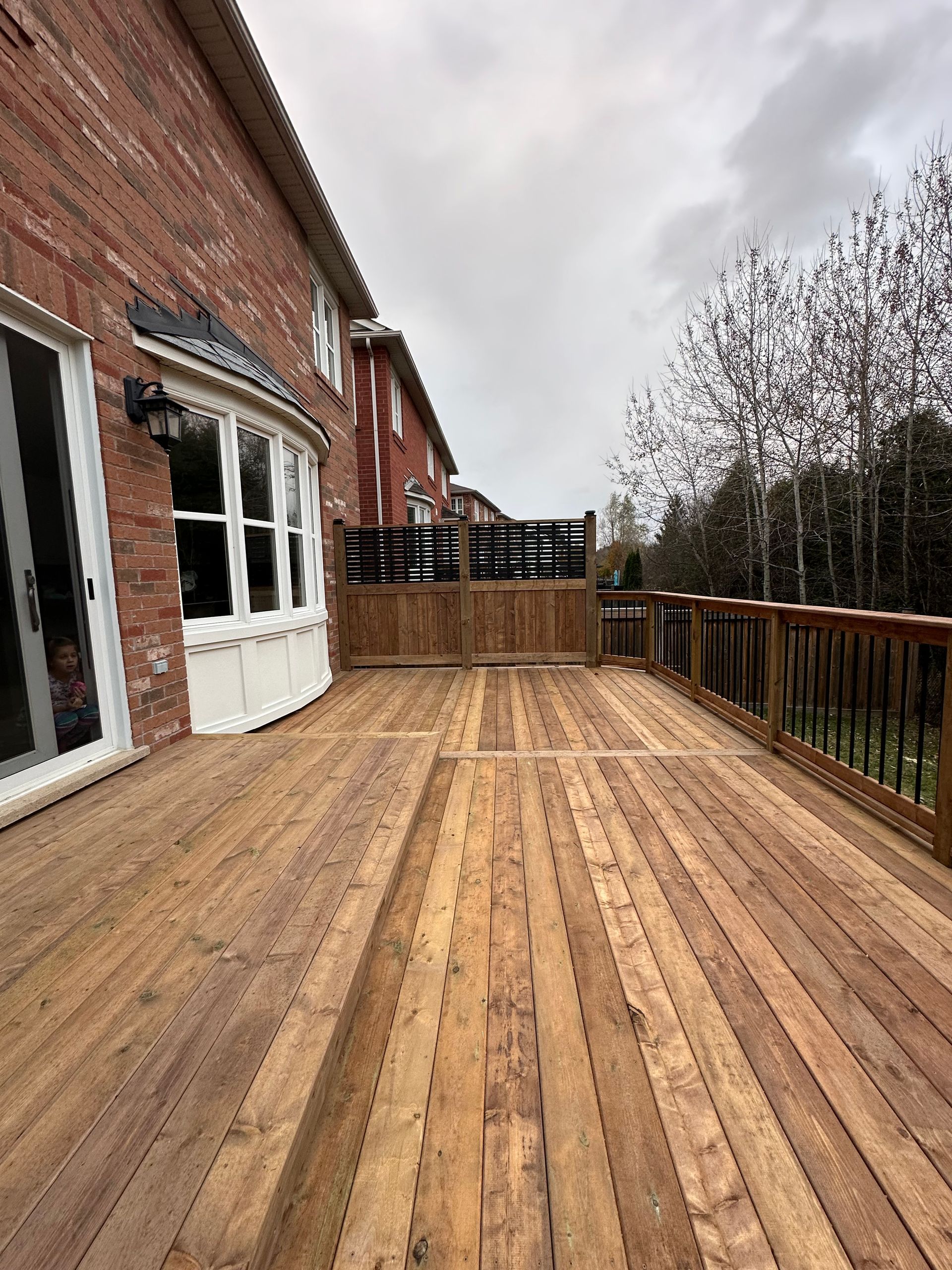 Wooden deck extends from brick house, railing along edge, overlooking backyard under cloudy sky.