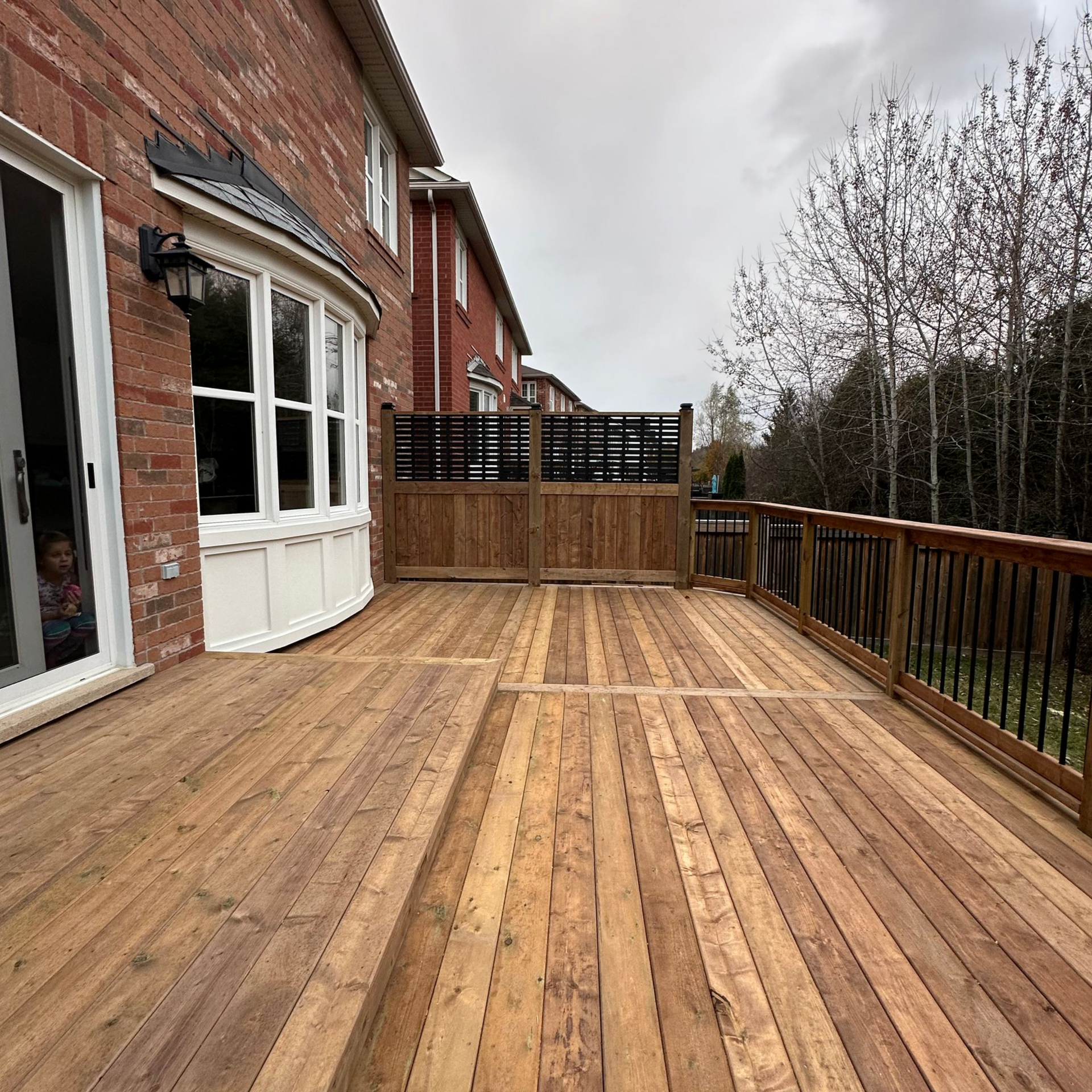 Wooden deck outside a brick house, partially fenced, with trees in the background.