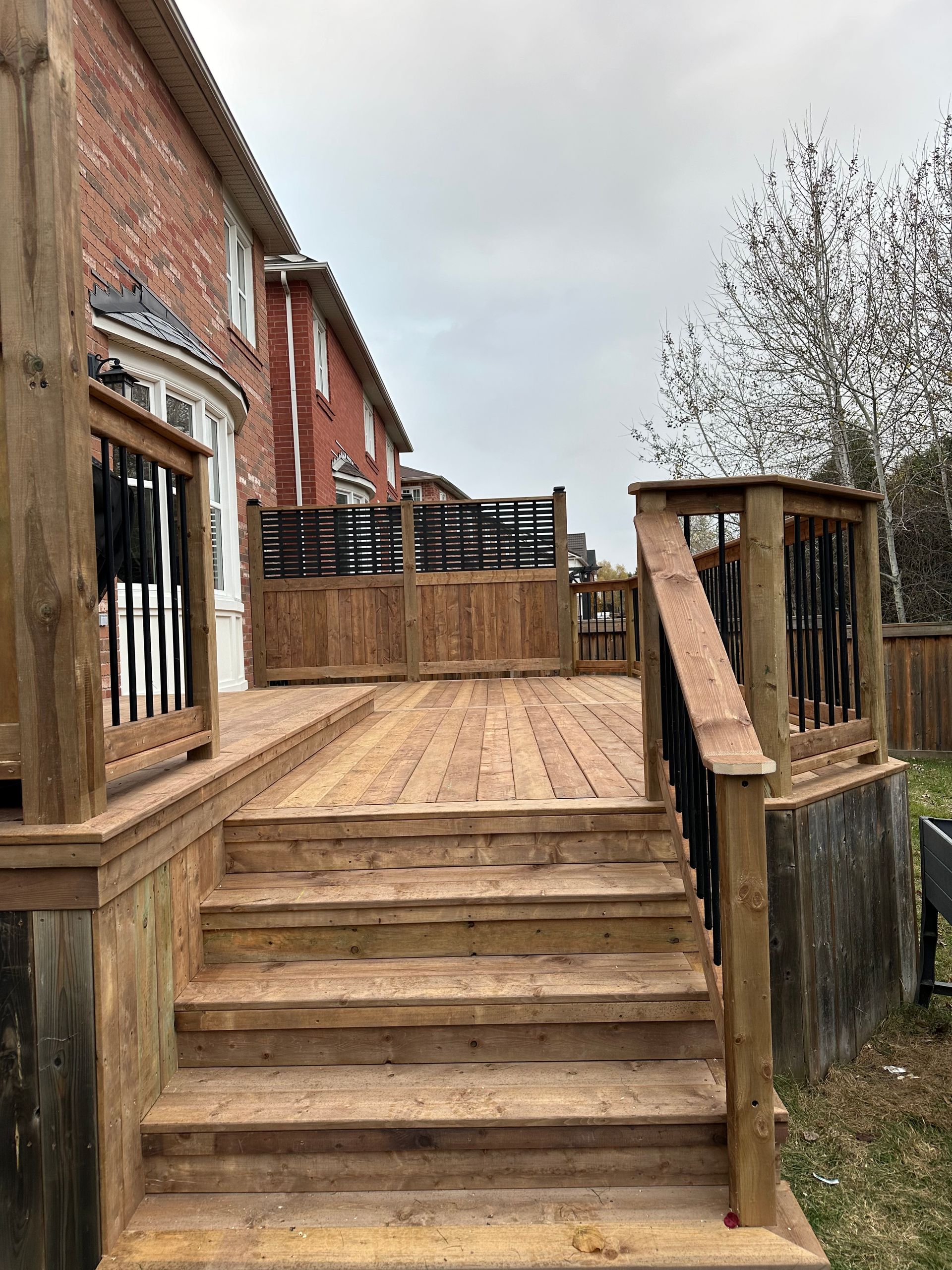 Wooden deck with stairs leading up to a house. Dark railings and a brick exterior. Overcast day.