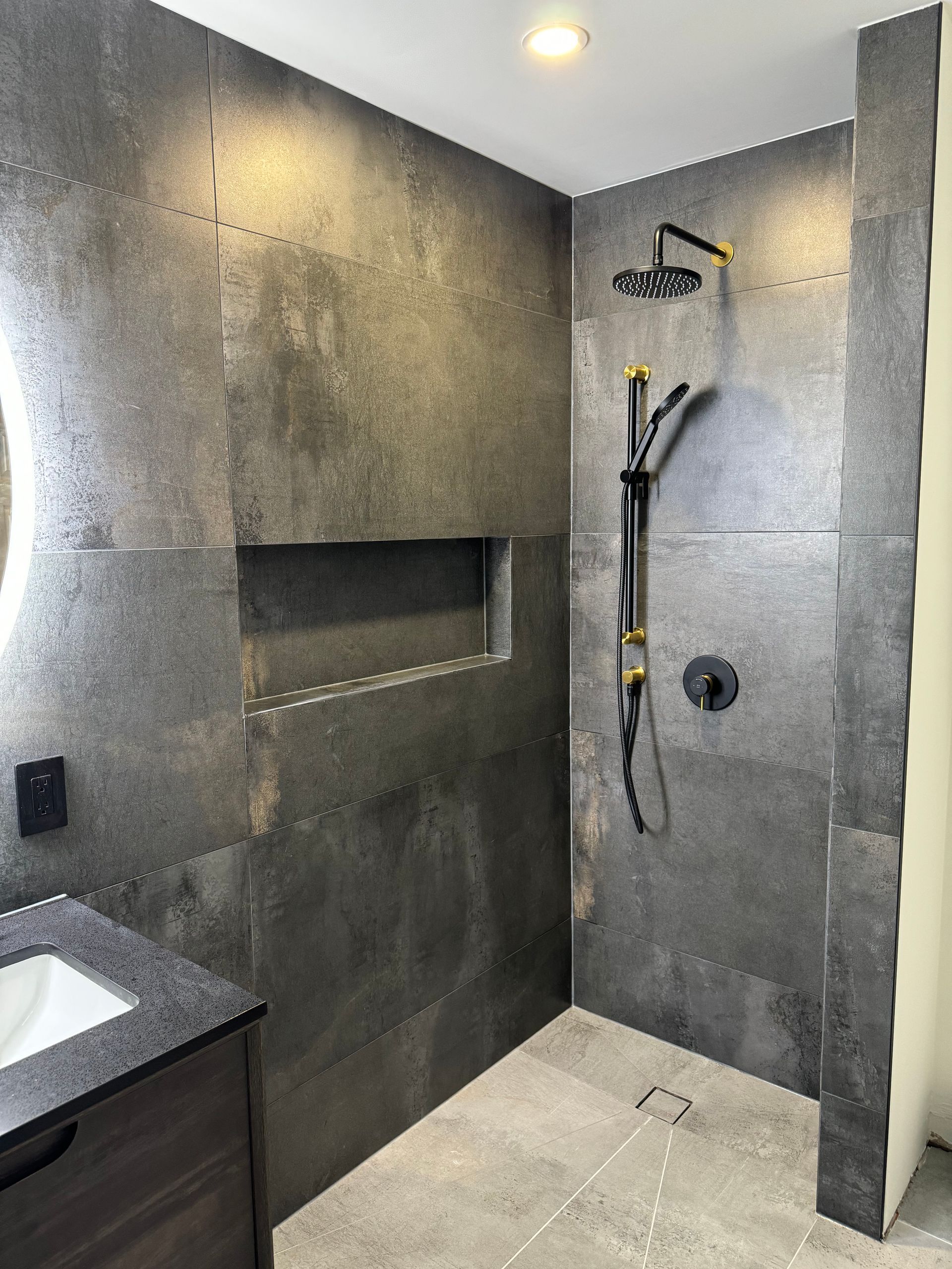 Dark gray tiled shower with black and gold fixtures, recessed shelf, and a dark vanity.