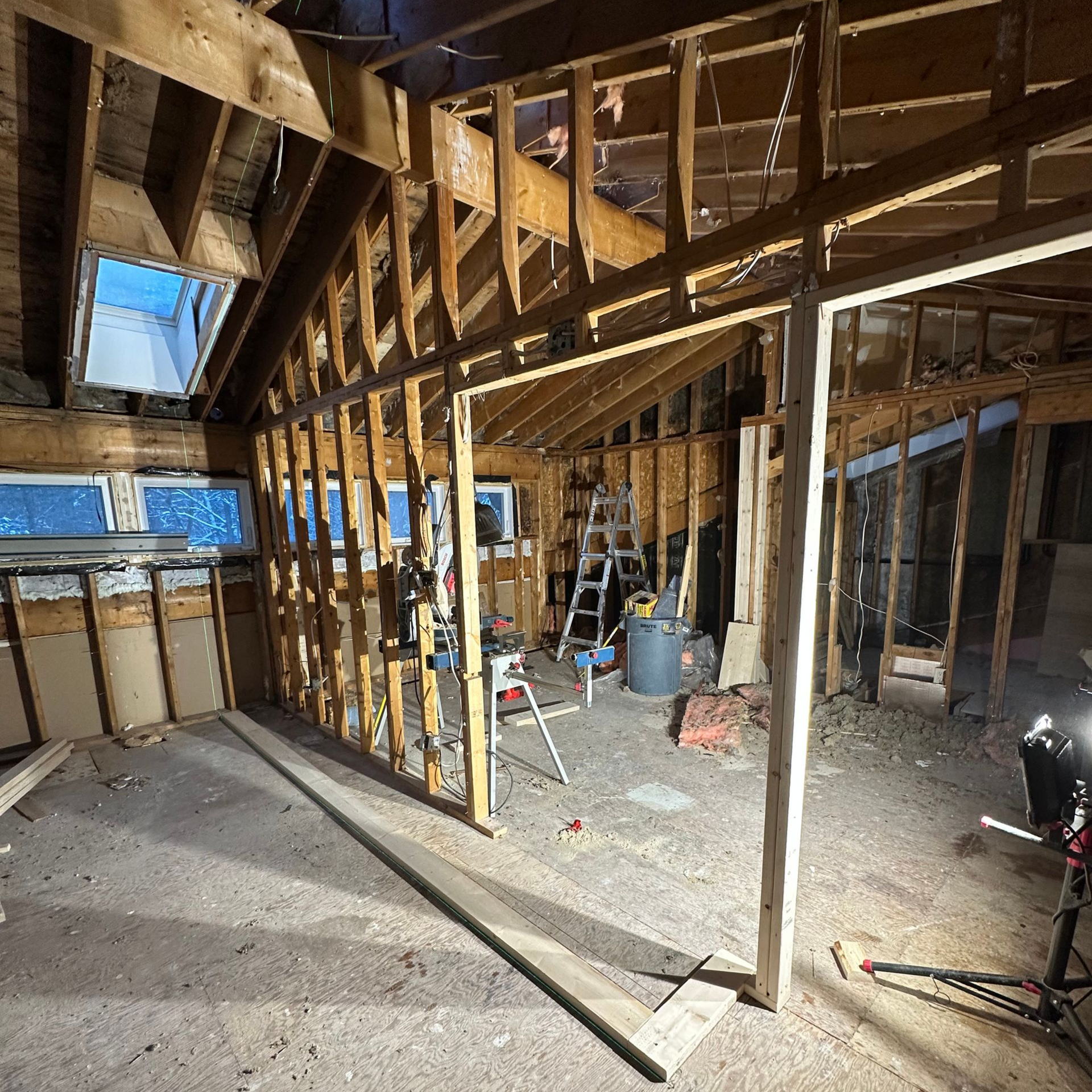 Interior view of a room under construction with exposed wooden framing and partial walls. Tools, windows, and sunlight visible.