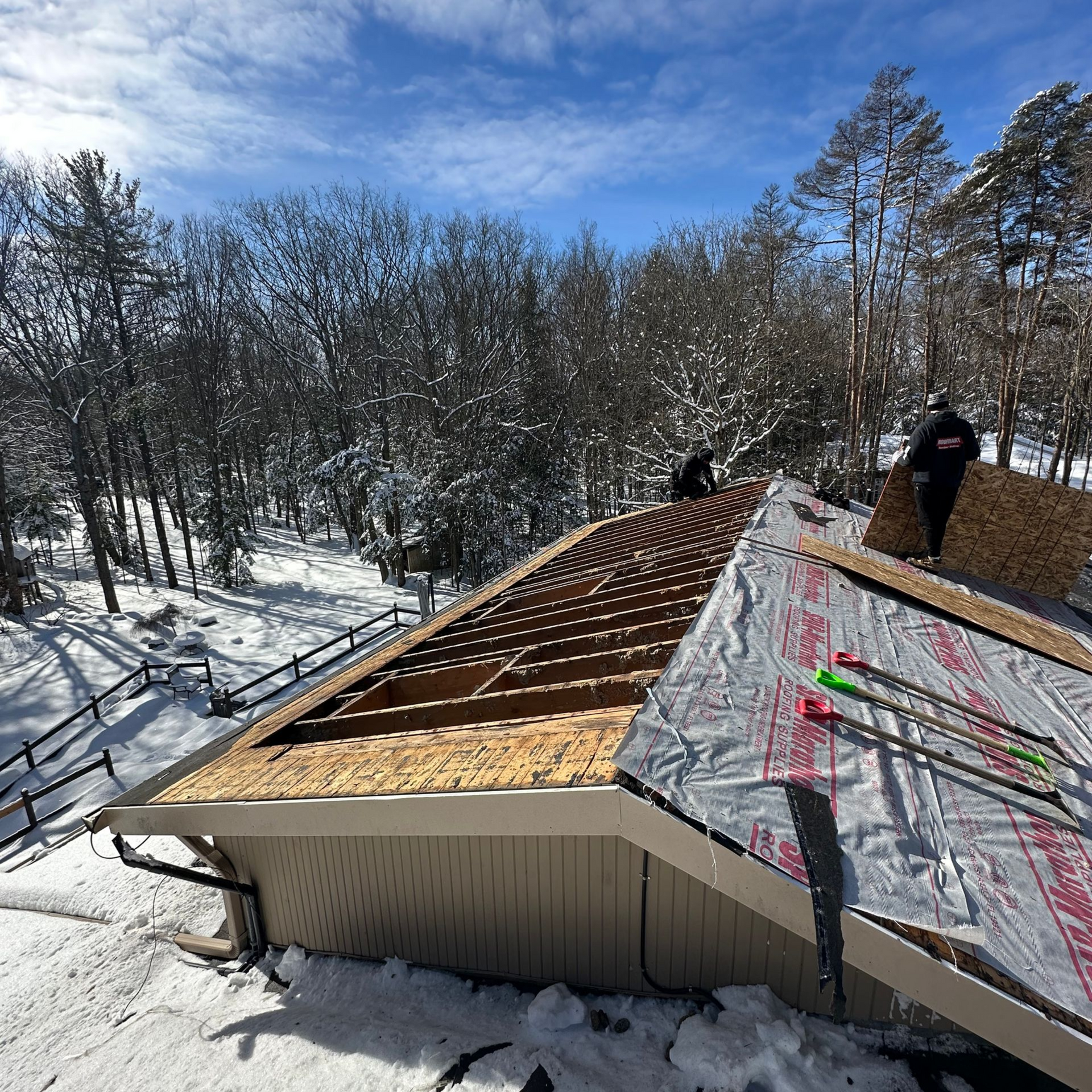 Roofers on a snowy roof installing shingles; trees and a blue sky in the background.