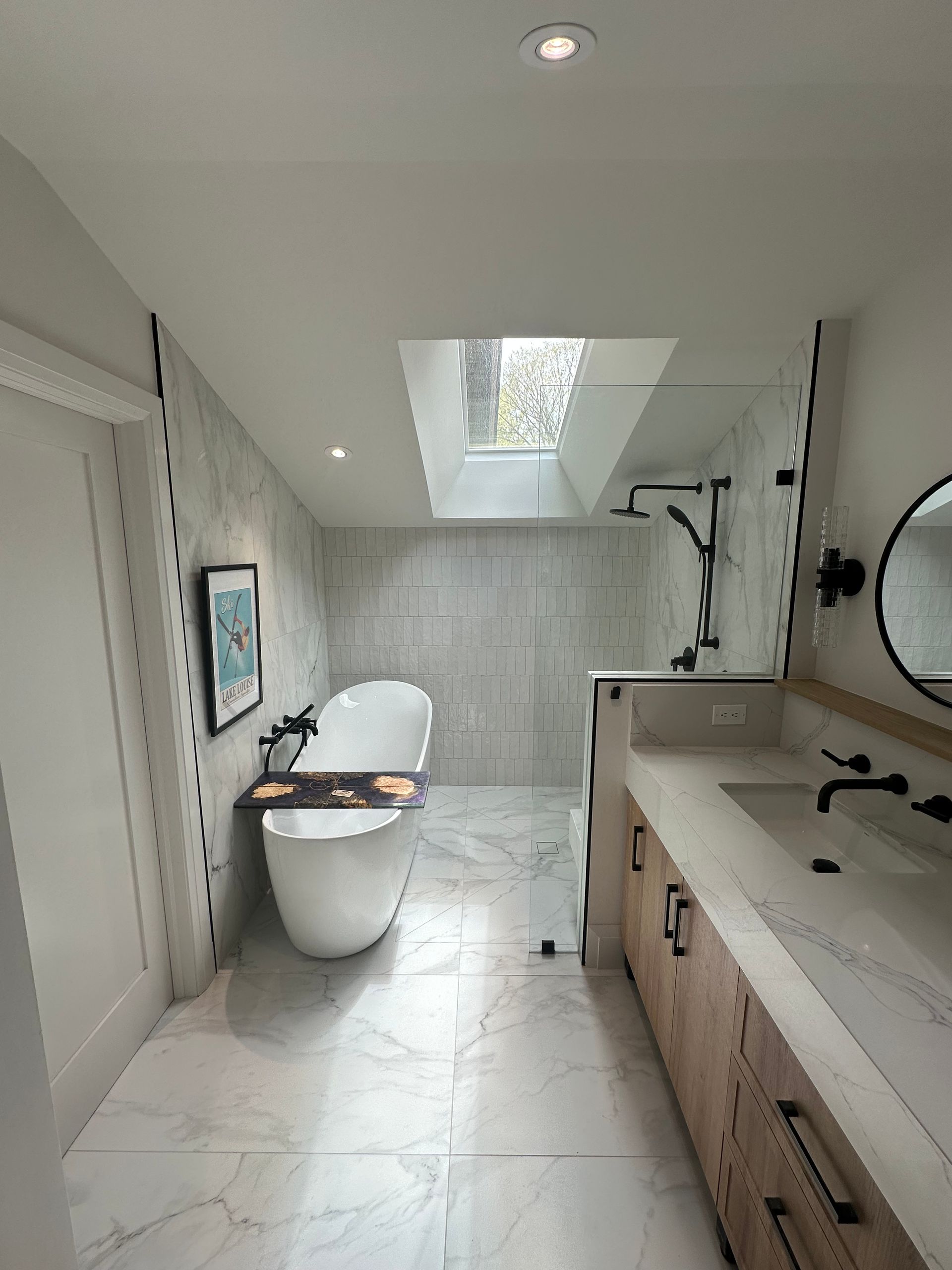 Modern bathroom with freestanding tub, marble tile, skylight, and double vanity.
