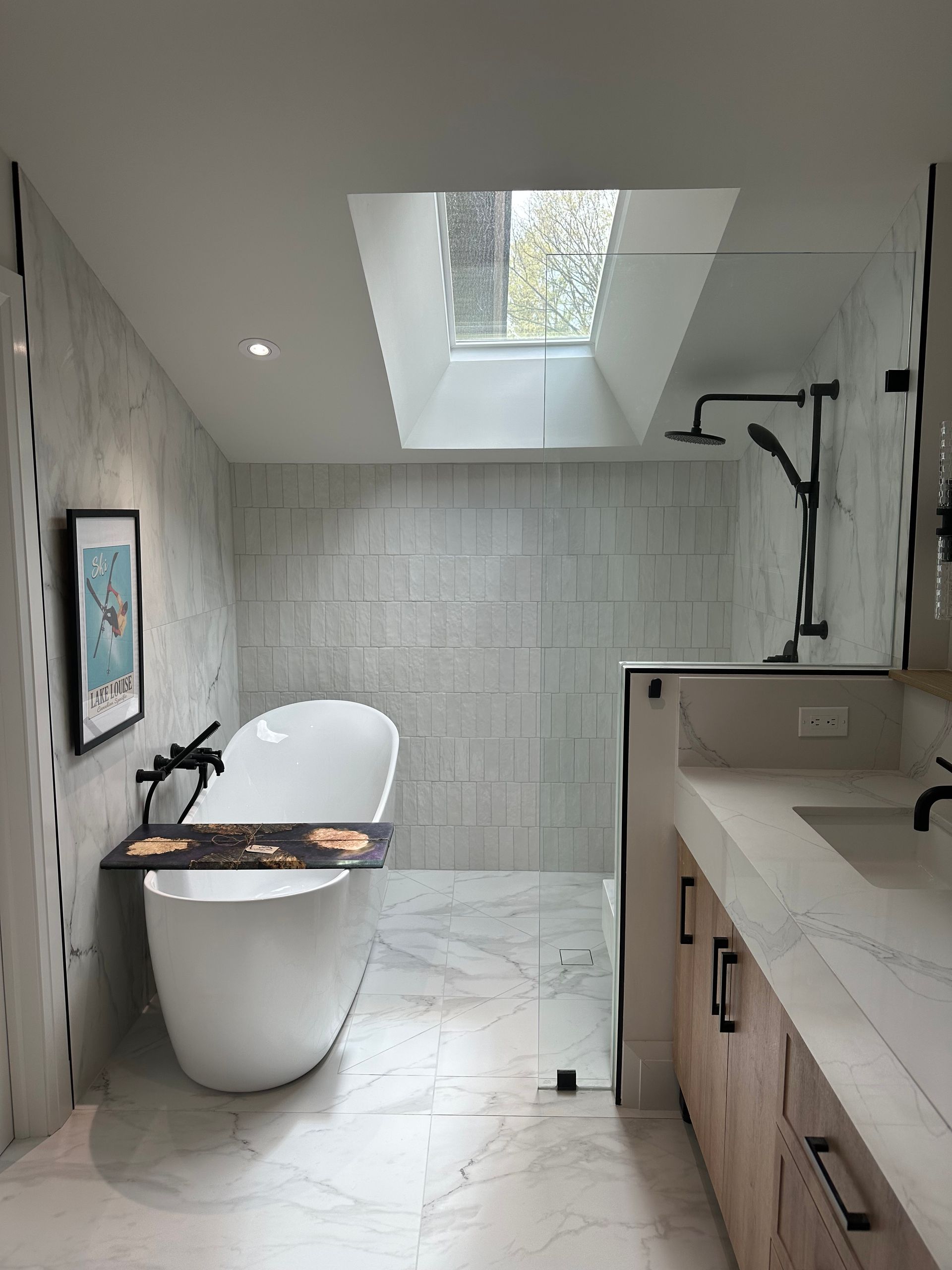 Modern bathroom with a white tub, shower, and vanity. Gray and white marble-look tiles. Skylight overhead.