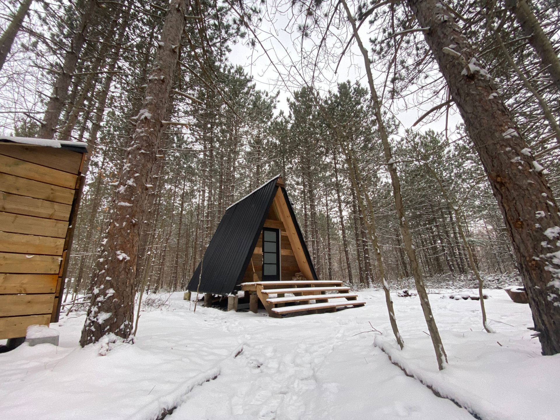 Snowy A-frame cabin in a forest, black roof, wooden stairs.