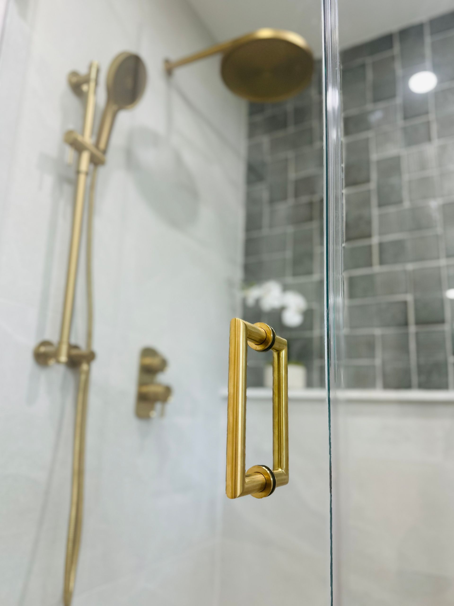 Gold shower fixtures in a modern bathroom with glass door, gray tile, and white orchid.