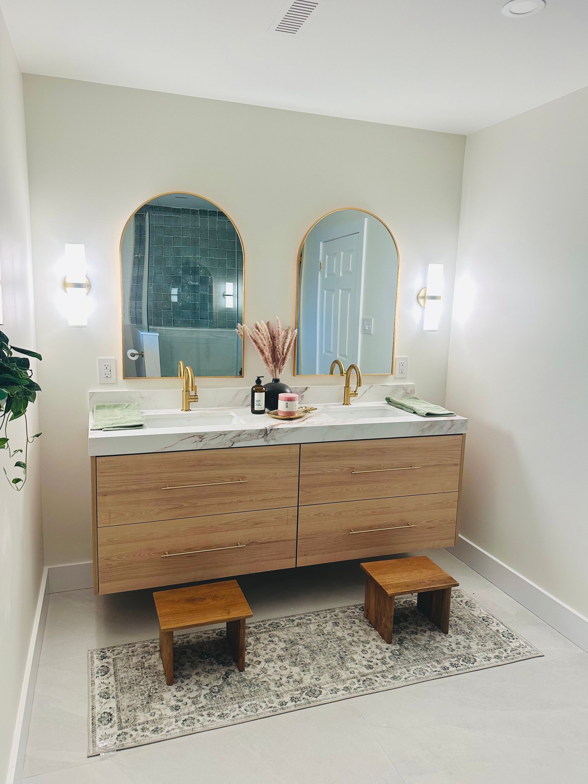 Bathroom with light wood double vanity, arched mirrors, gold fixtures, and two small stools on a patterned rug.