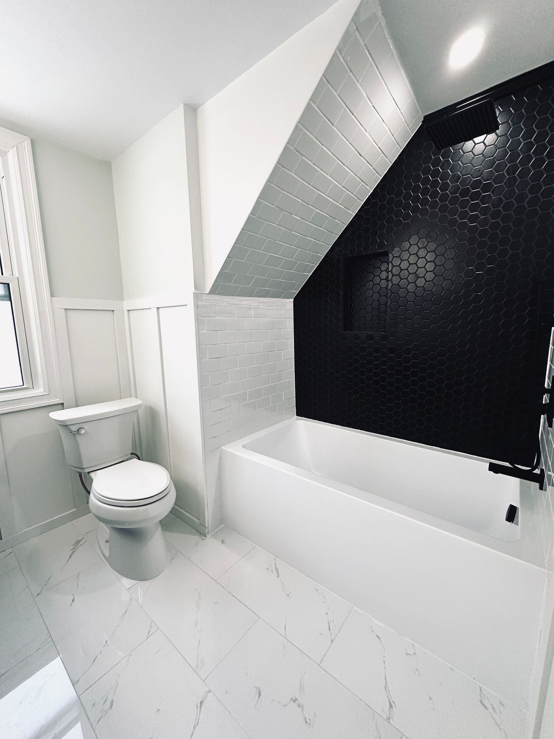 Modern white and black bathroom with hexagon tile accent wall, tub, toilet, and window.