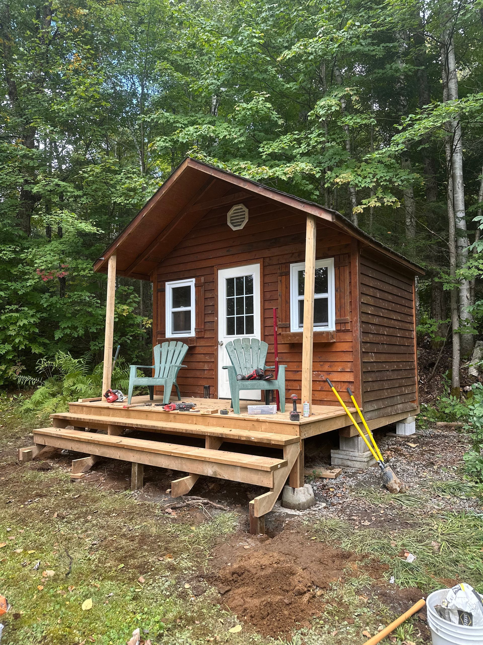 Small brown cabin with porch, two green chairs, set in a forest.