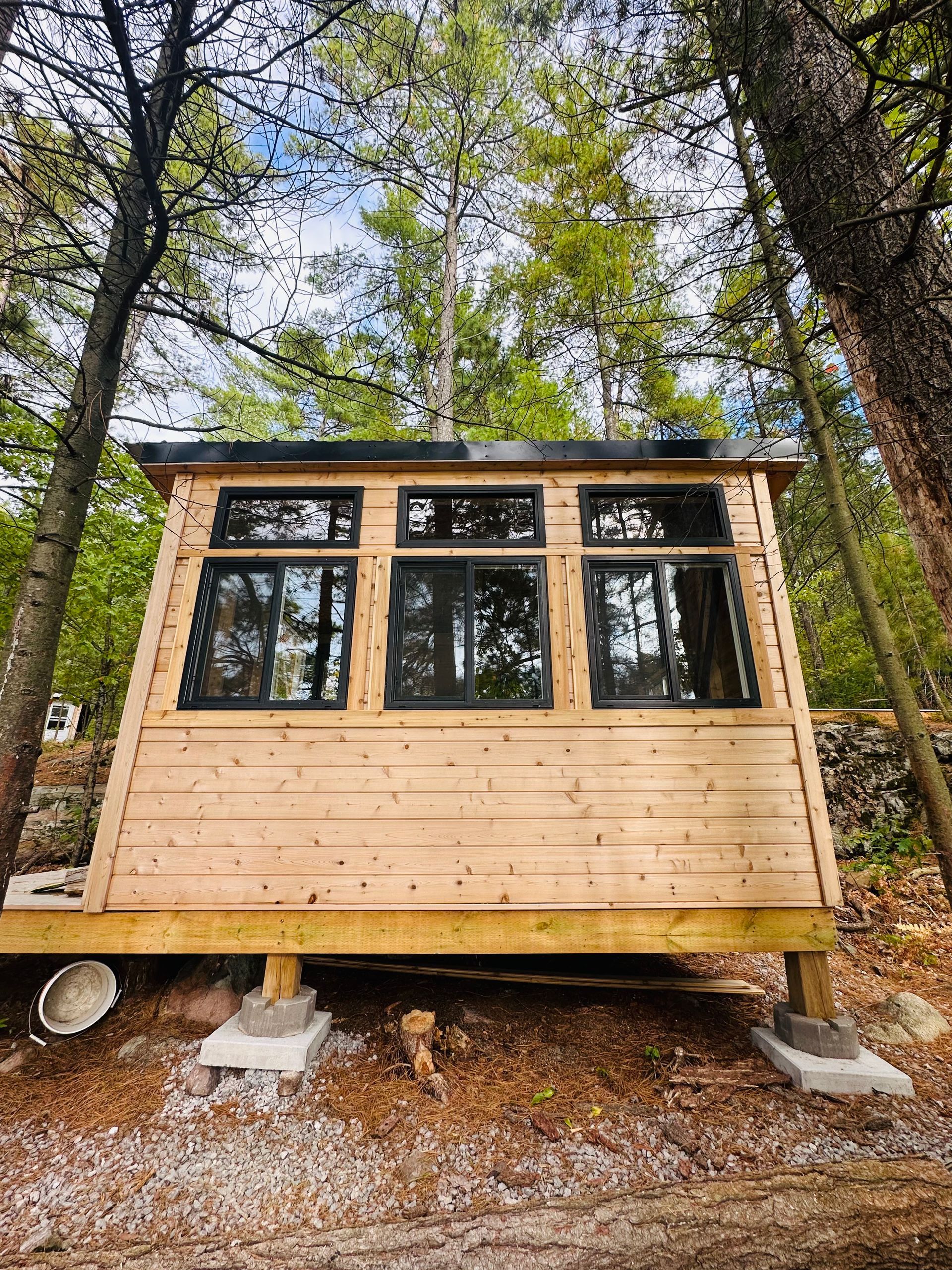 Wooden cabin with black framed windows in a forest, elevated on concrete blocks.